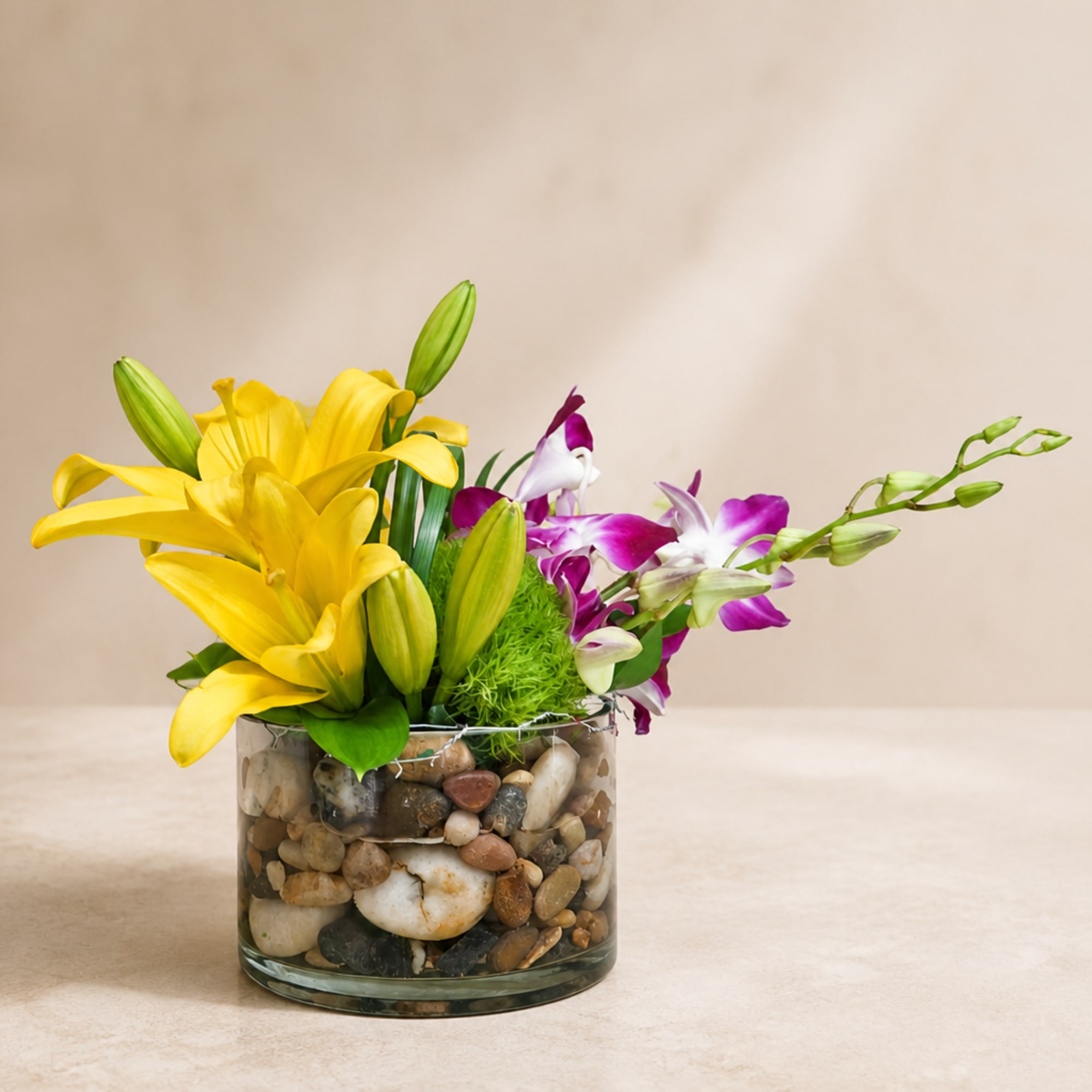 Yellow lilies and purple orchids in a pebble-filled glass vase