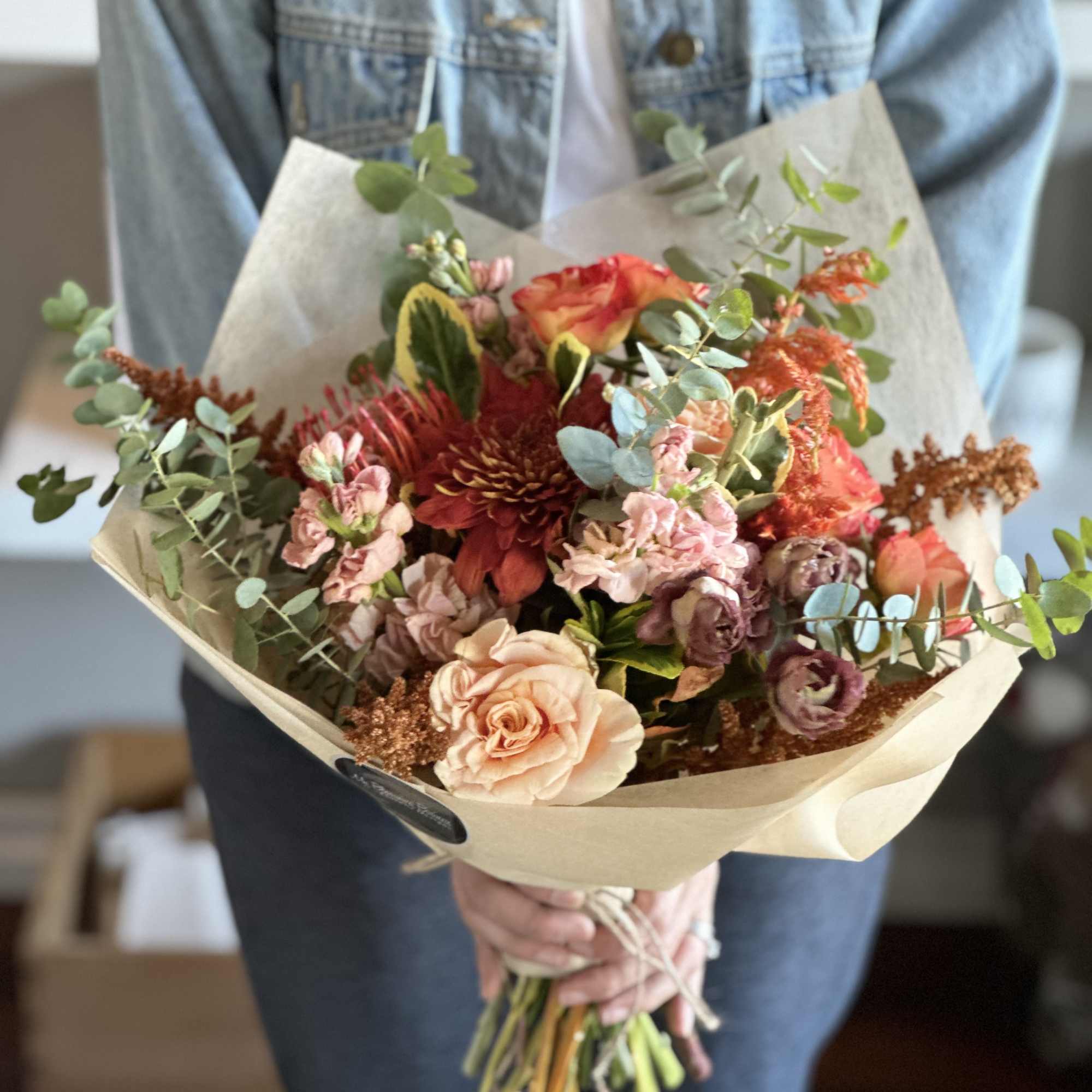 Handheld bouquet of peach, red, and pink flowers wrapped in paper