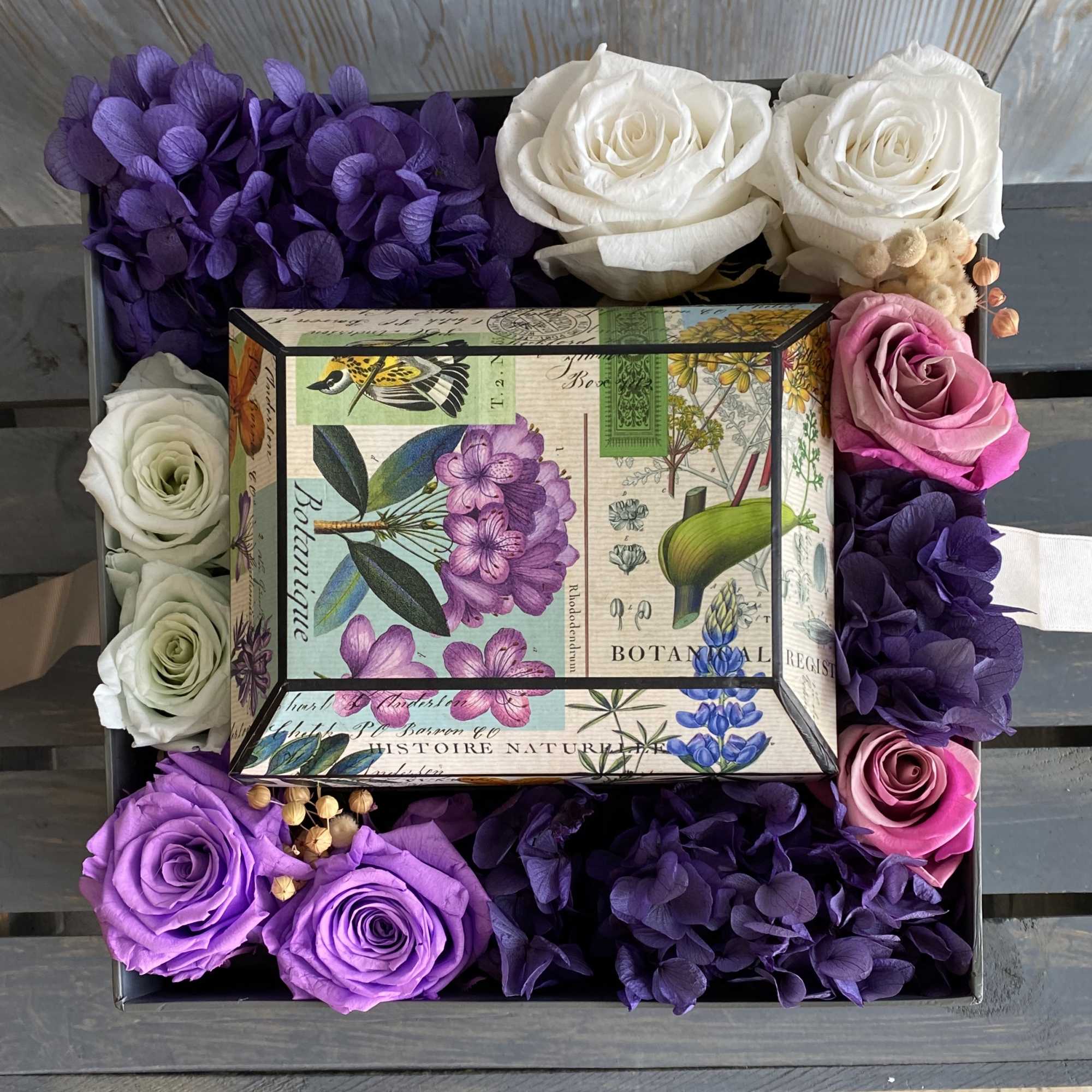 Boxed arrangement of purple, pink, and white roses with hydrangea petals