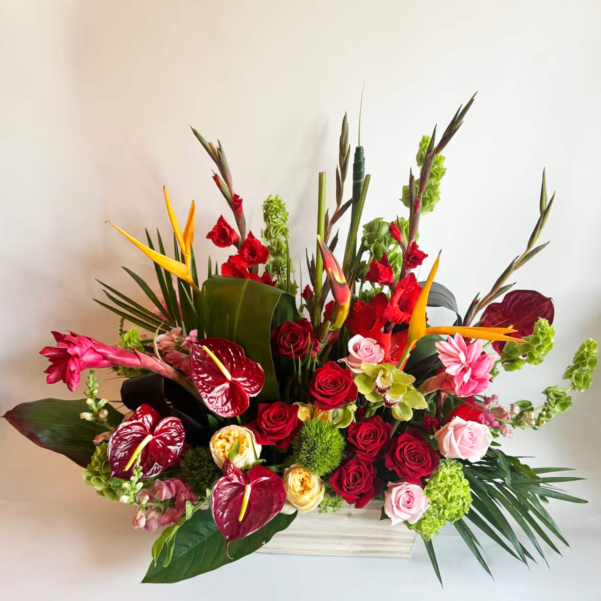 Tropical floral arrangement with red roses, pink anthuriums, and orange bird-of-paradise in a white container.