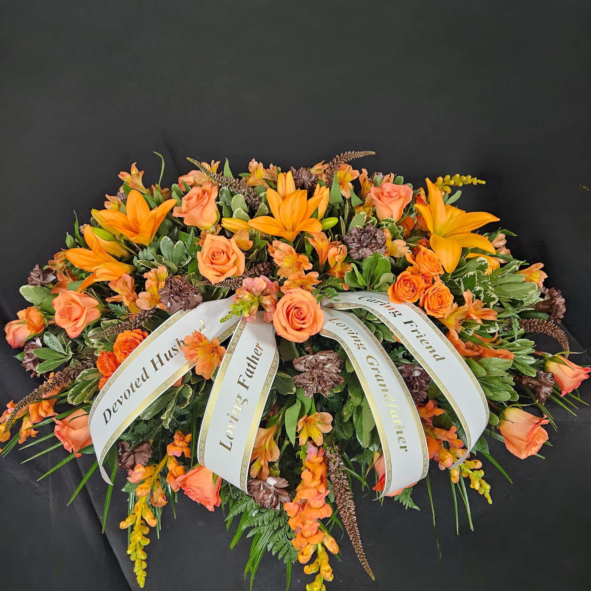 Orange lilies and roses casket spray with white sympathy ribbons on a dark background