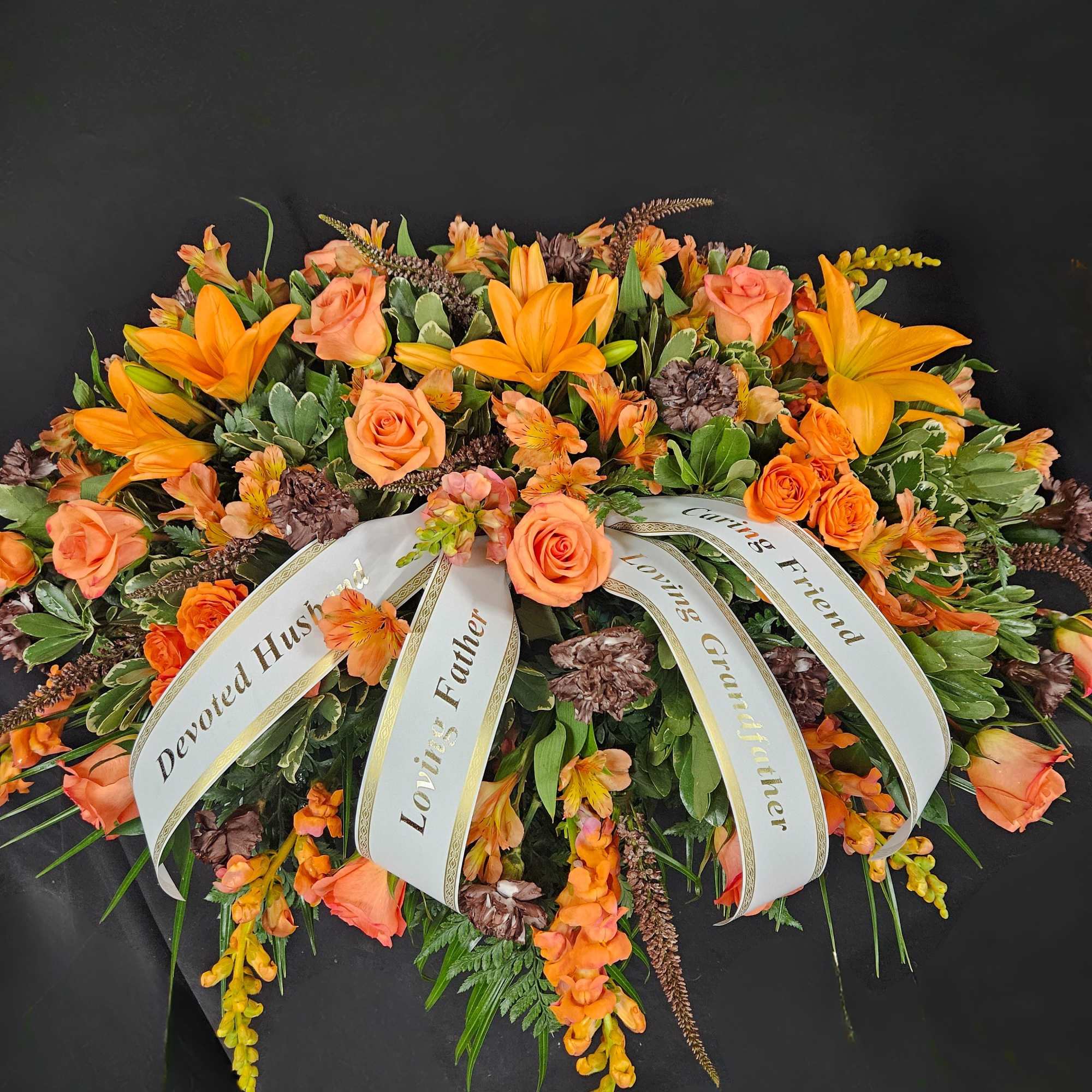 Sympathy casket spray with orange lilies, roses, and memorial ribbons on a black background