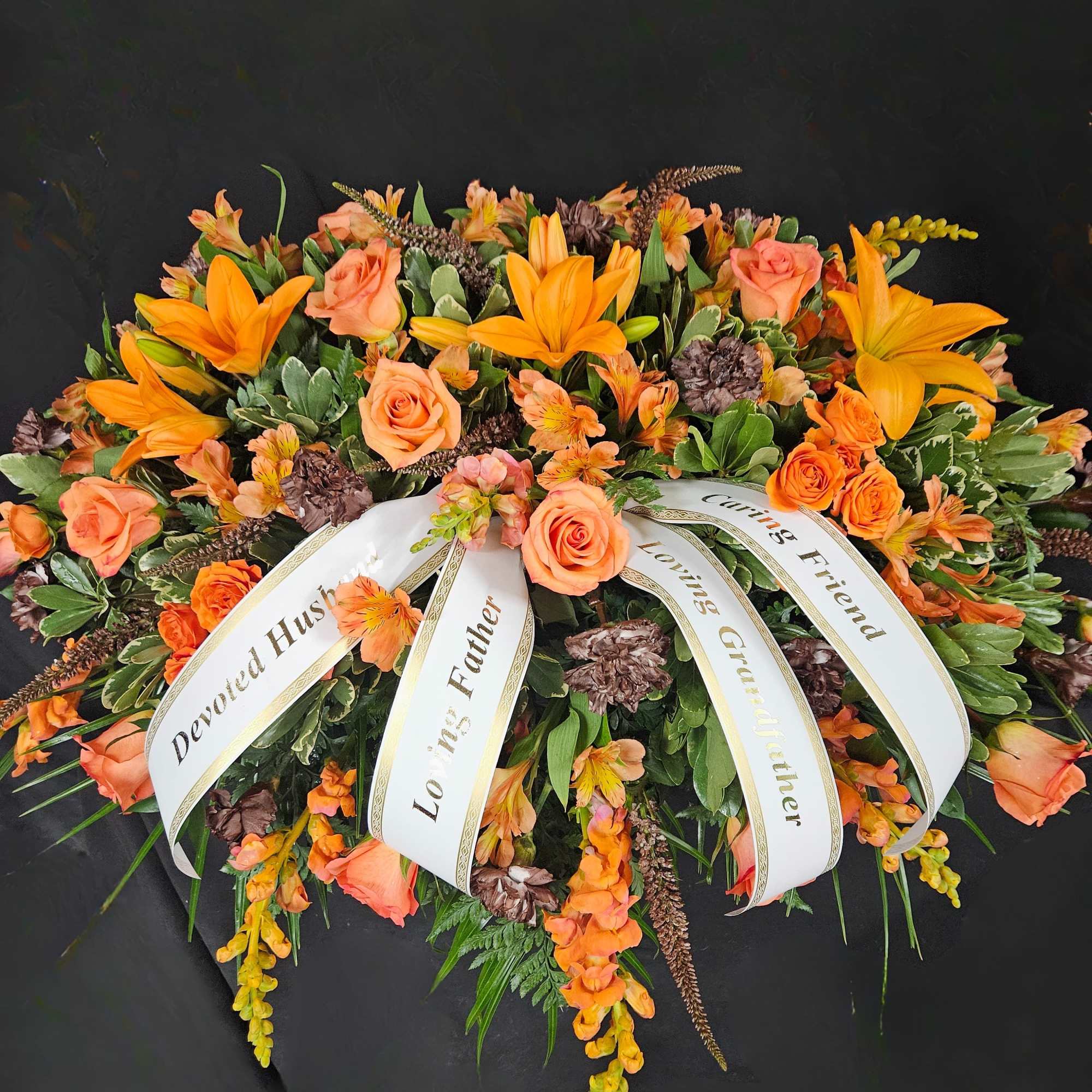 Elongated orange lily and rose funeral spray with white memorial ribbons on a dark background.