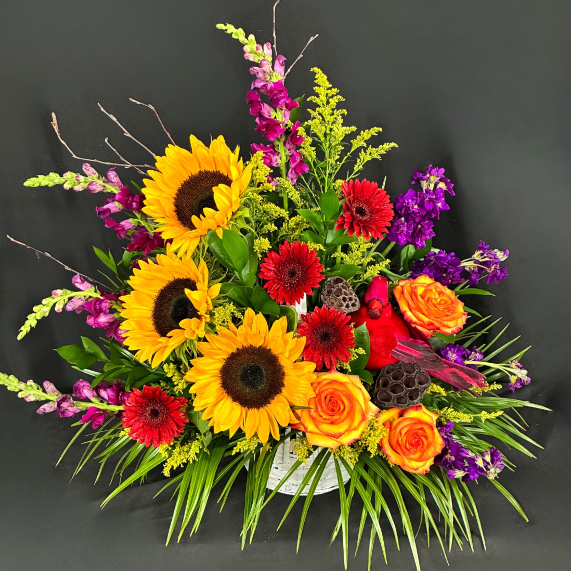 Colorful flower arrangement with sunflowers, roses, and red daisies in a white basket