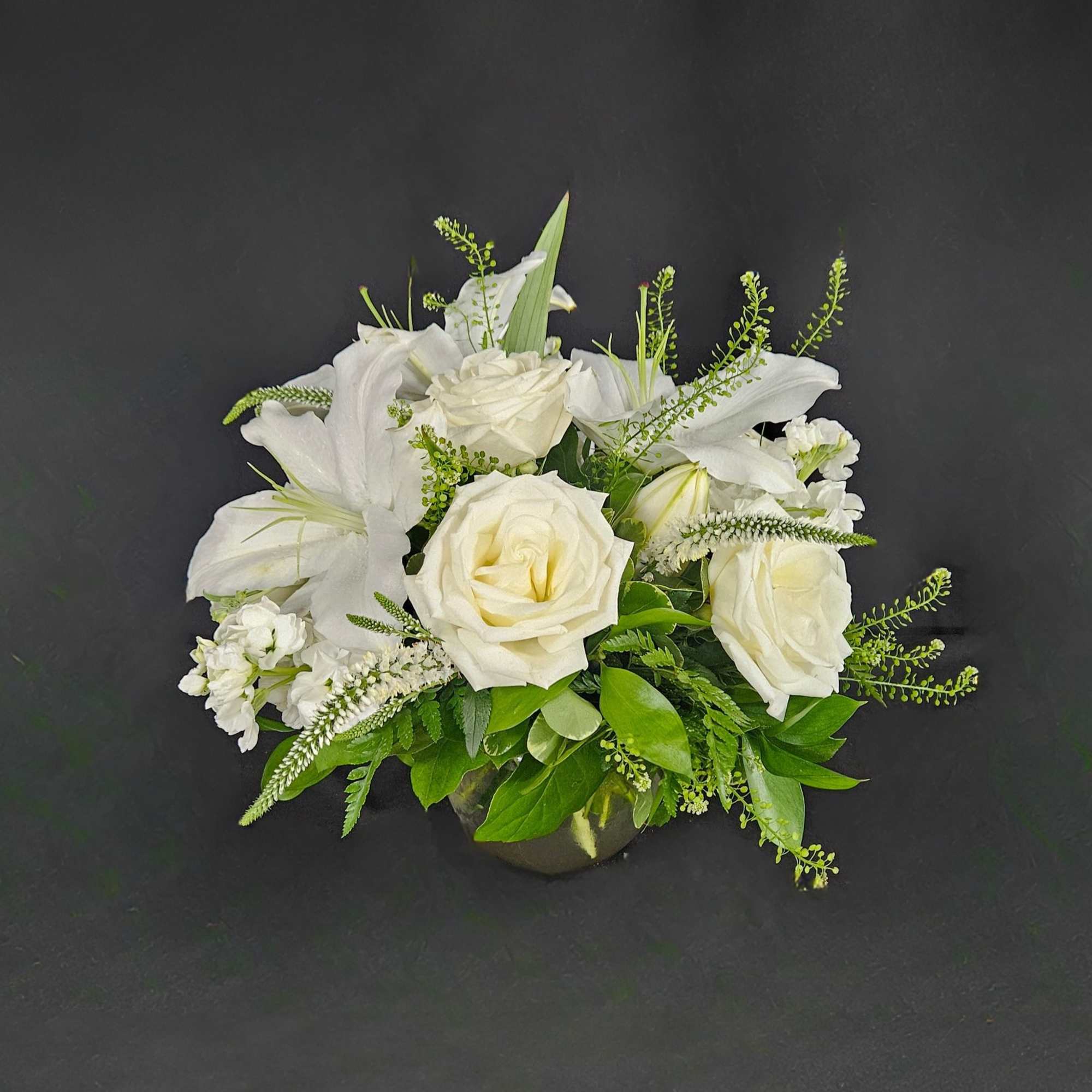 This arrangement includes white roses, white lilies, white stock, and dusty miller.