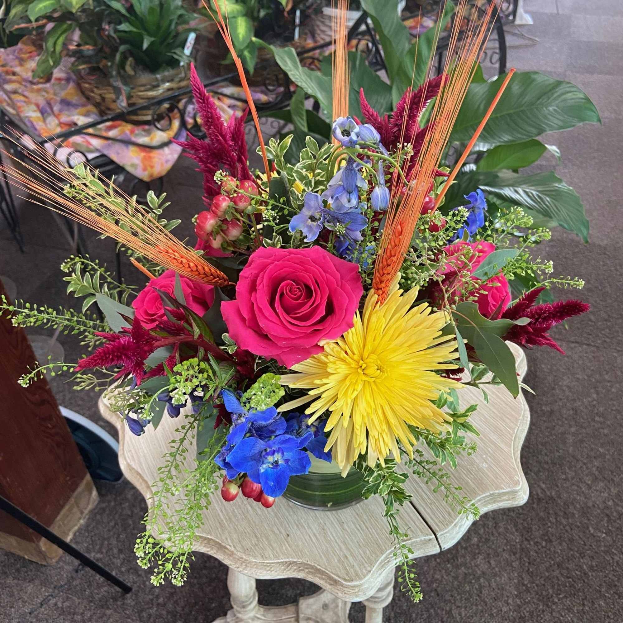 Bright mixed arrangement with hot pink rose, yellow mum, blue delphinium and wheat stems in a clear glass vase.