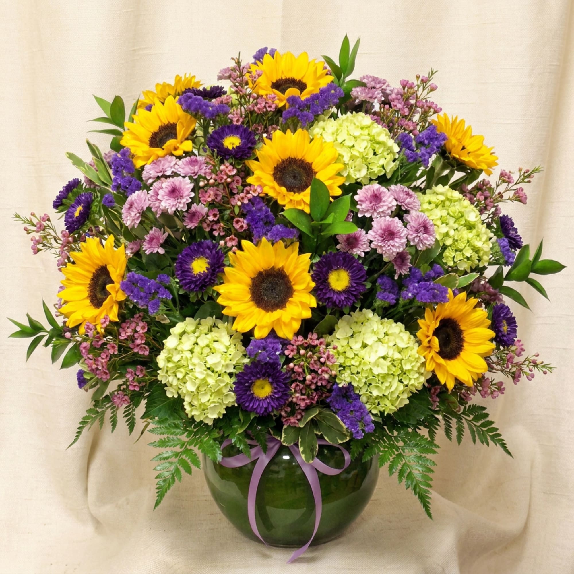 Sunflowers and purple flowers arranged in a green glass vase with a ribbon.