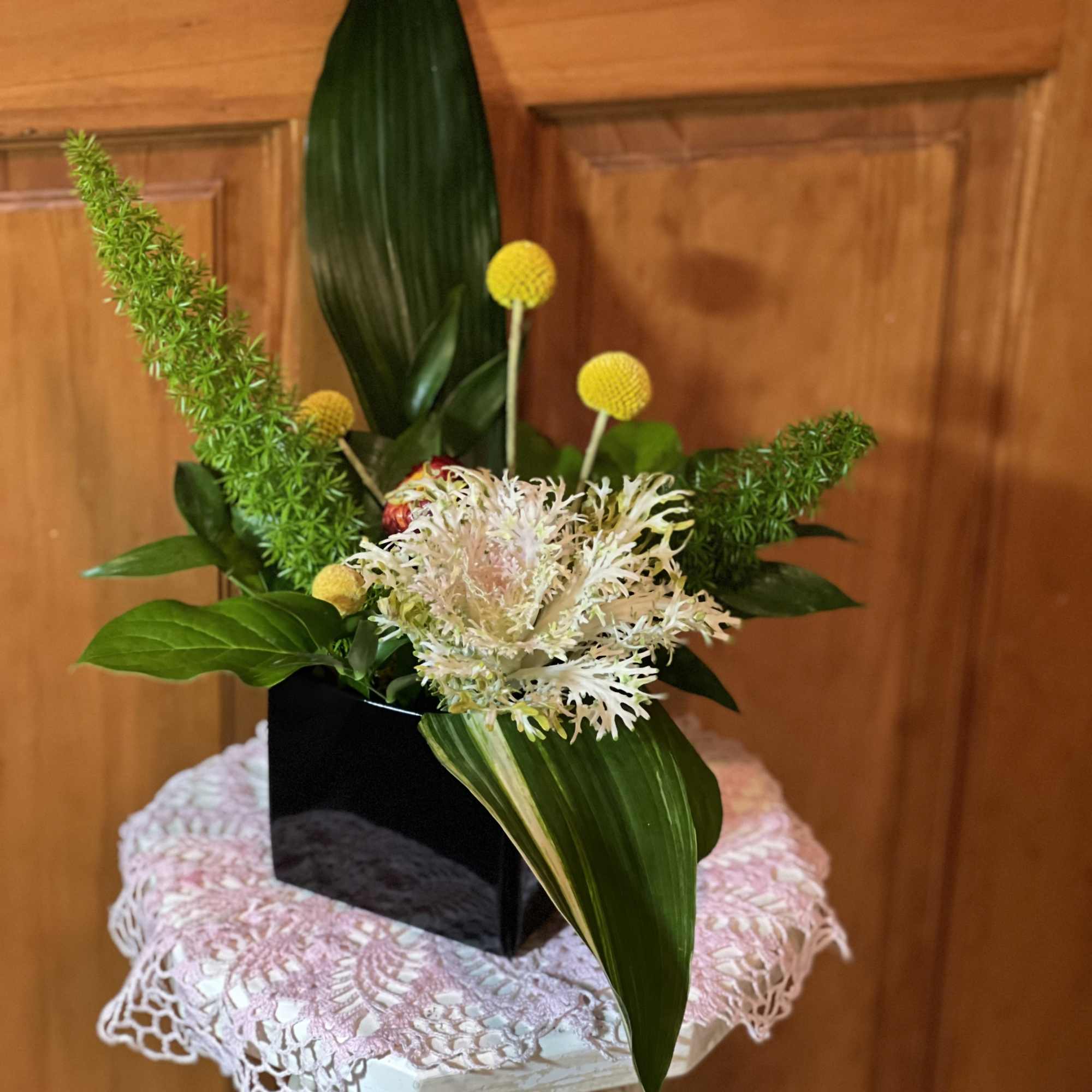 Compact floral arrangement with white blooms, yellow accents, and broad green leaves in a black vase