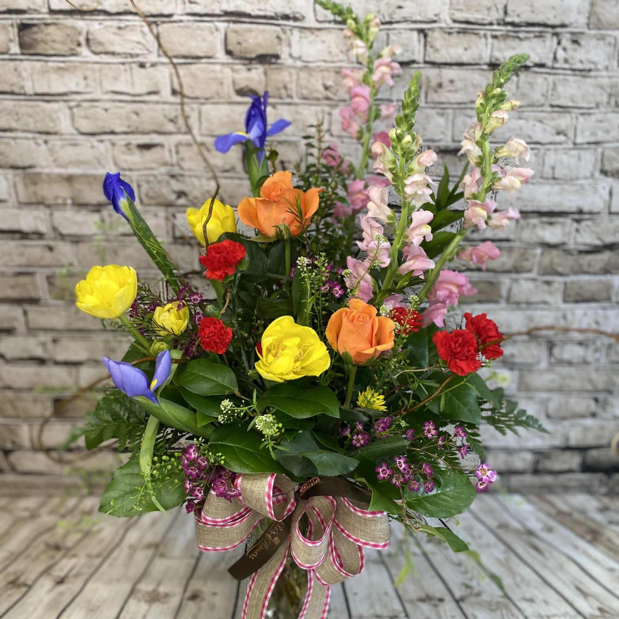 Tall mixed arrangement with yellow and orange roses, red carnations, blue iris and pink snapdragons in a glass vase