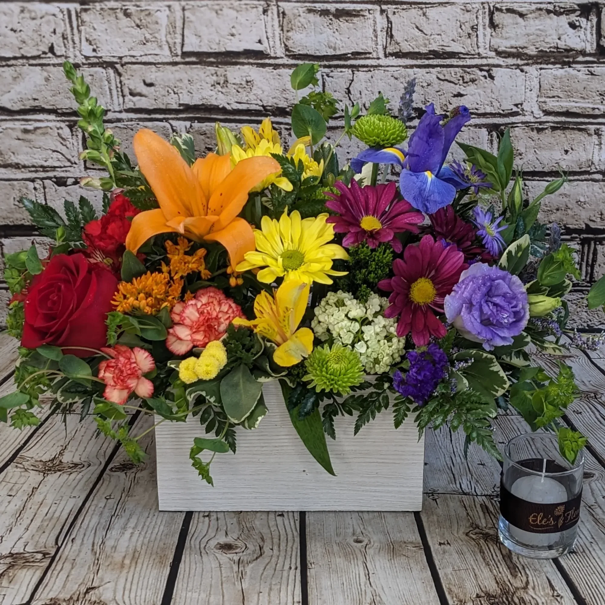 Colorful mixed flower arrangement in a white rectangular box with a glass candle holder