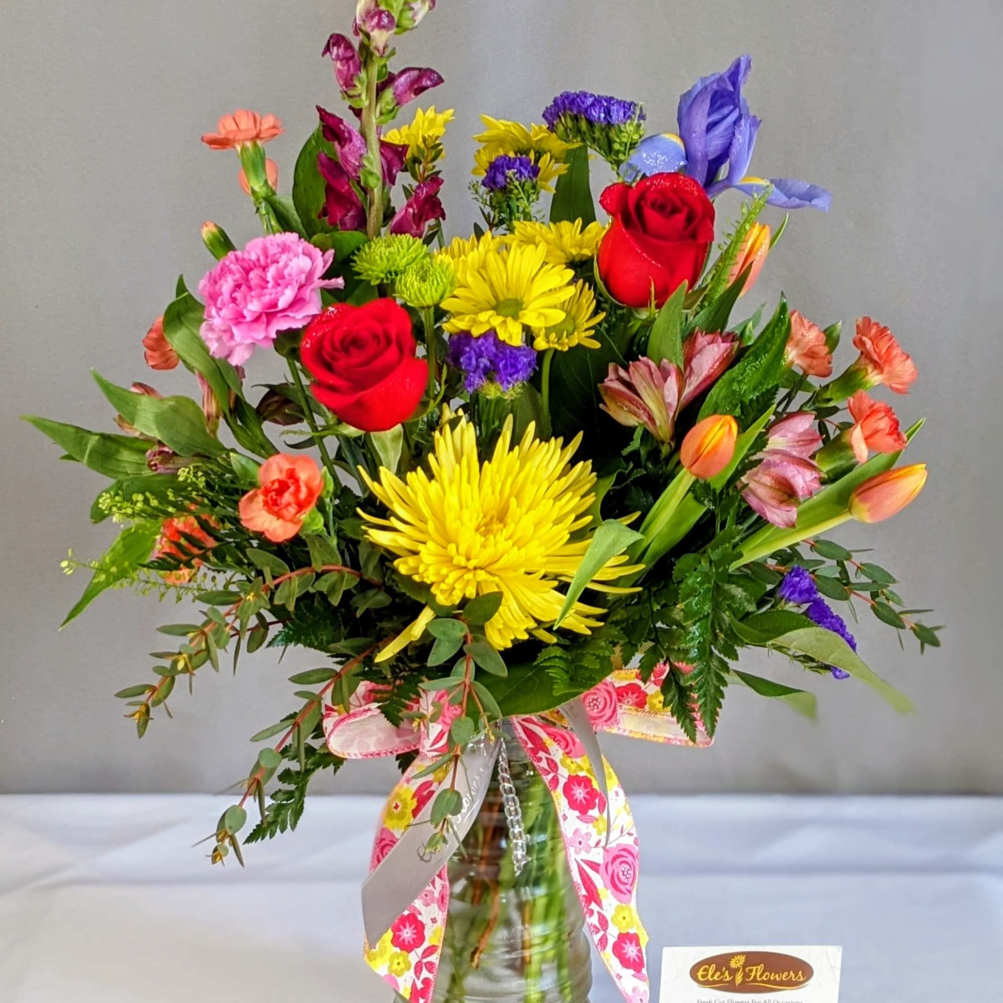 Tall mixed bouquet of bright roses, mums, tulips, and assorted flowers in a glass vase with a colorful ribbon.