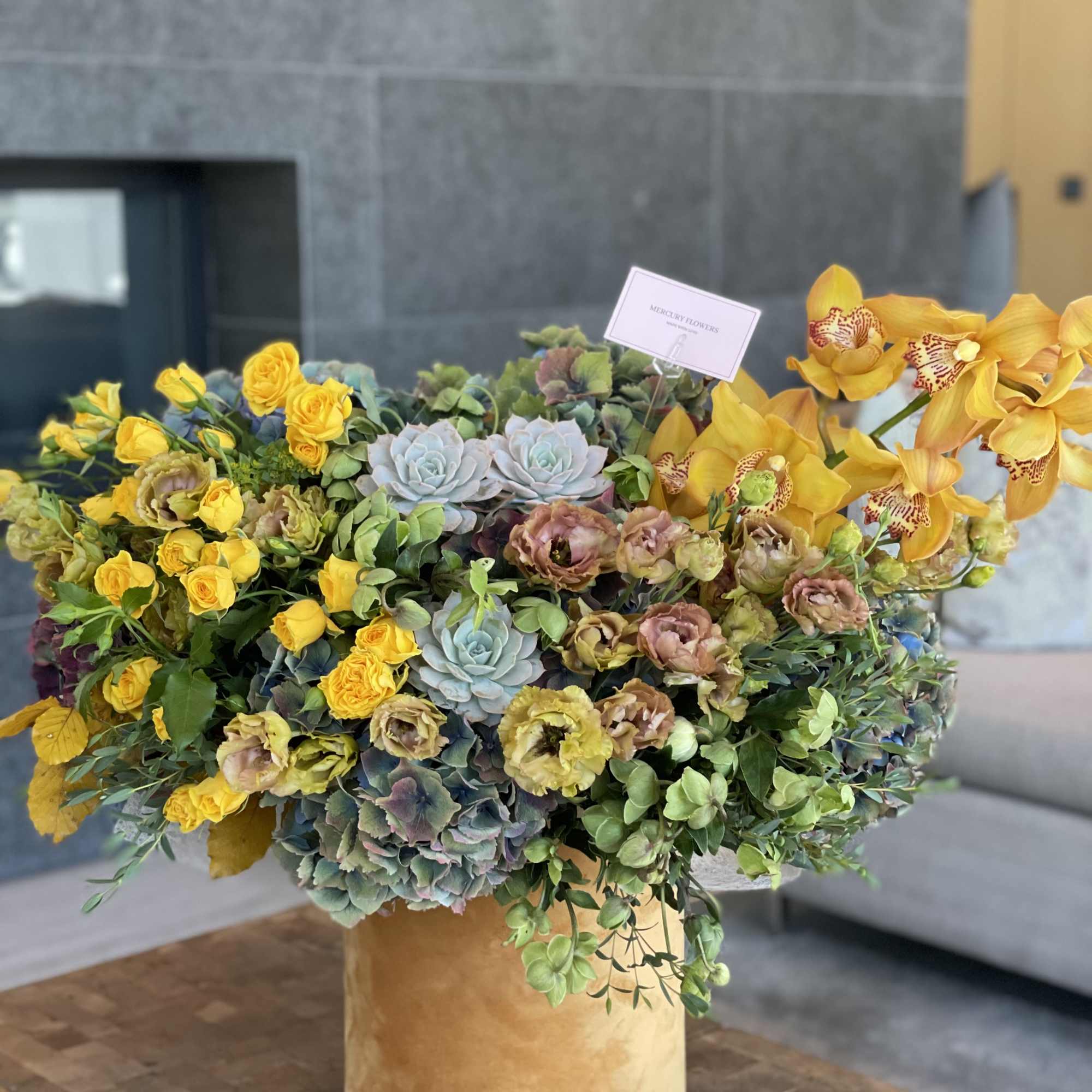 Large floral arrangement with yellow roses, orchids, and succulents in a gold vase.