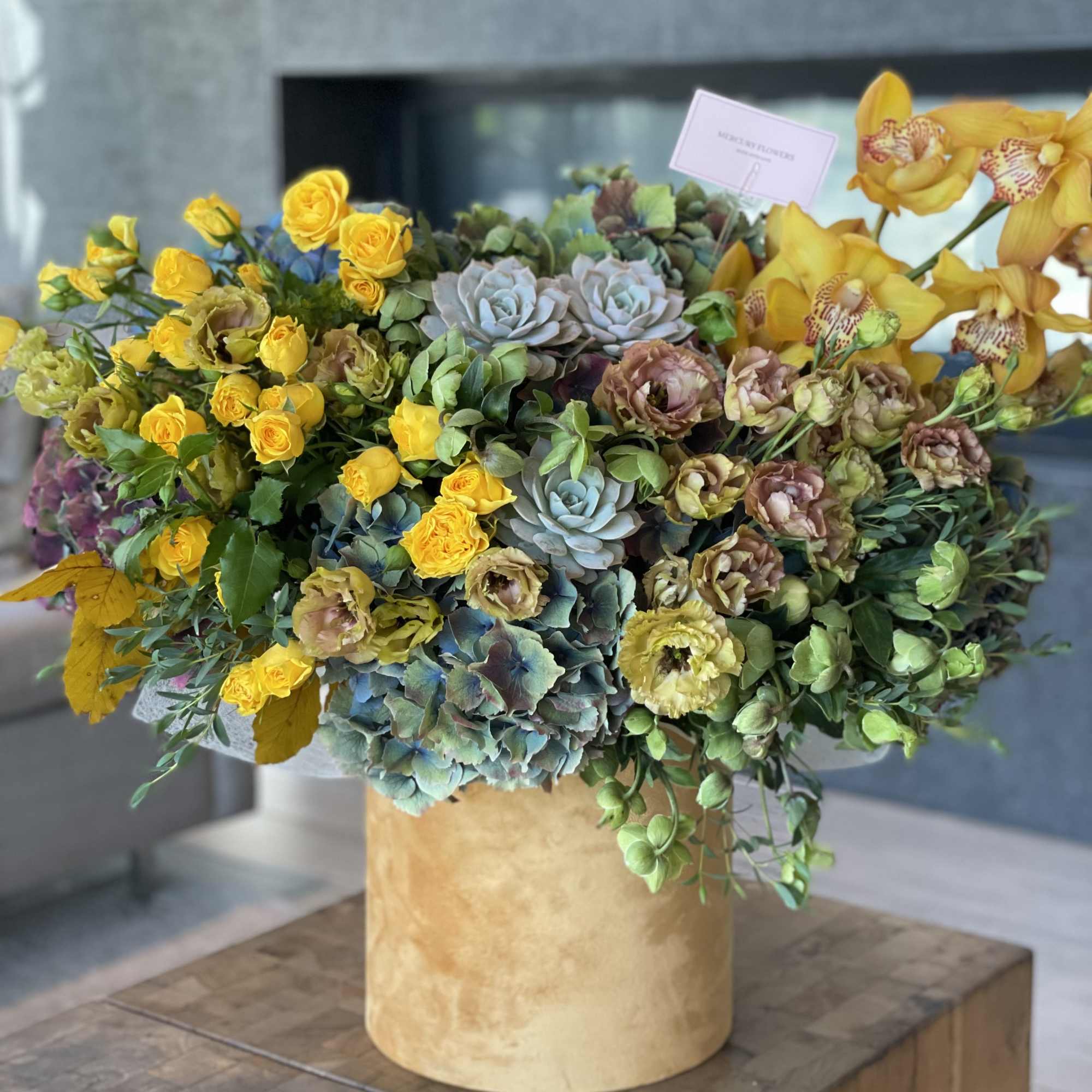 Large mixed floral arrangement in a gold vase with yellow roses and orchids