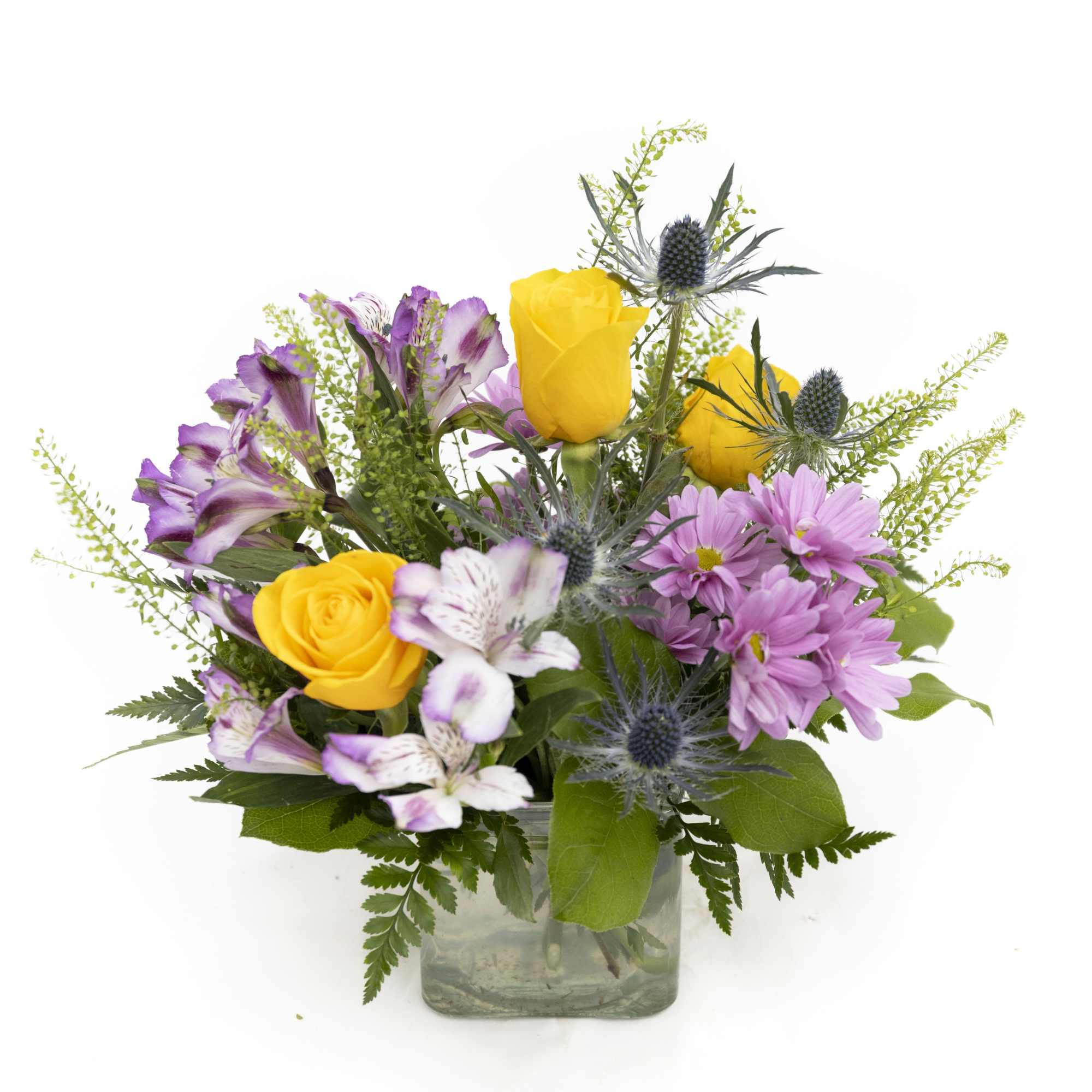 Yellow roses, purple daisies, alstroemeria, and blue thistles in a clear glass cube vase
