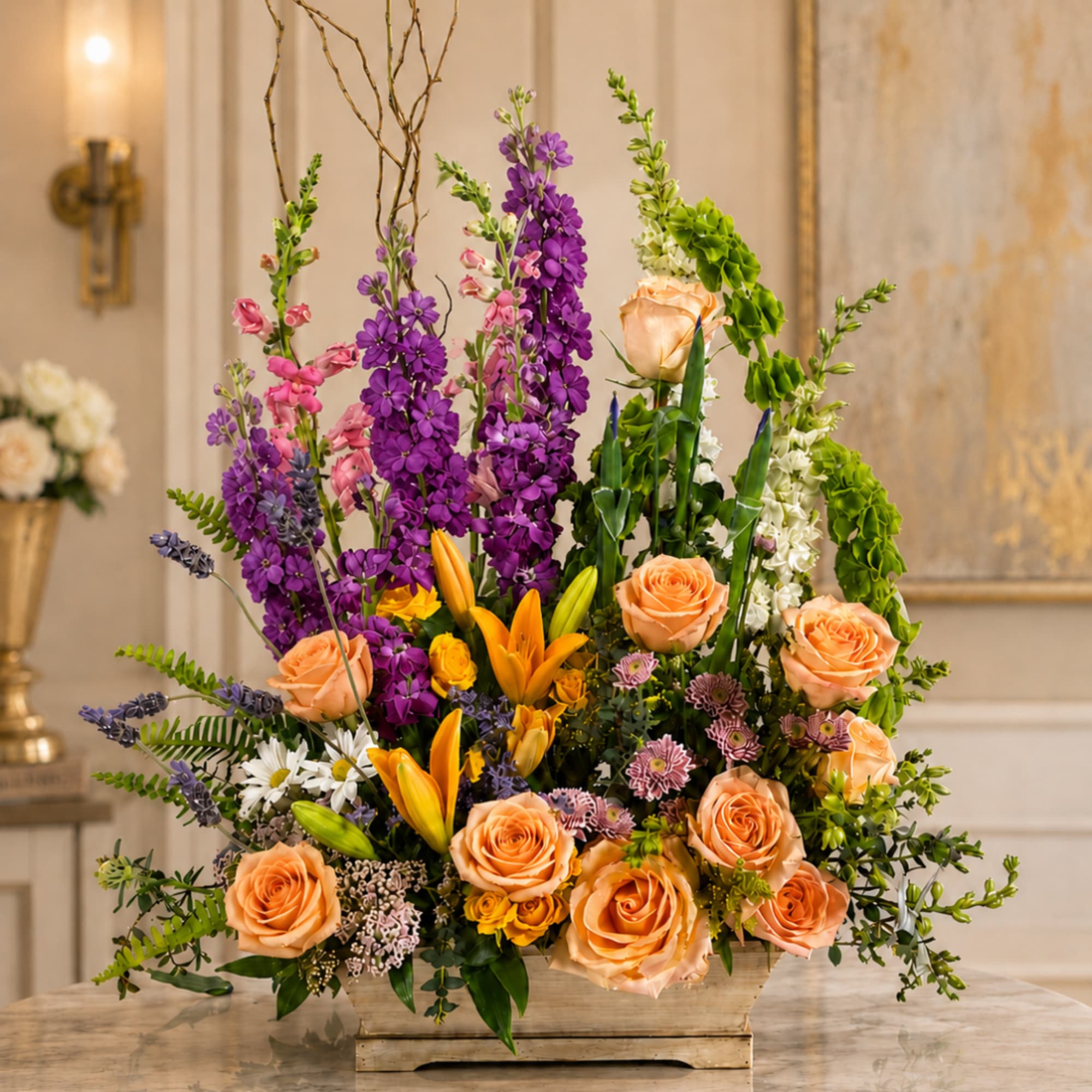 Tall mixed flower arrangement in a rectangular container with peach roses and purple blooms
