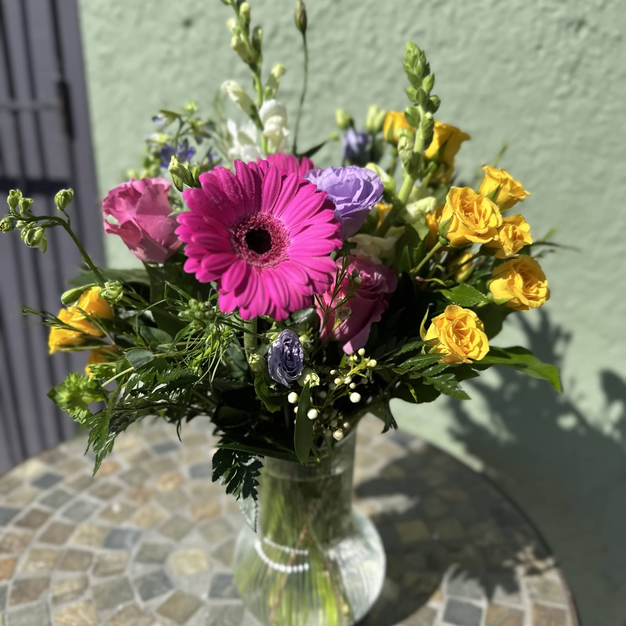 Mixed bouquet of pink gerbera, yellow roses, and purple blooms in a clear glass vase.