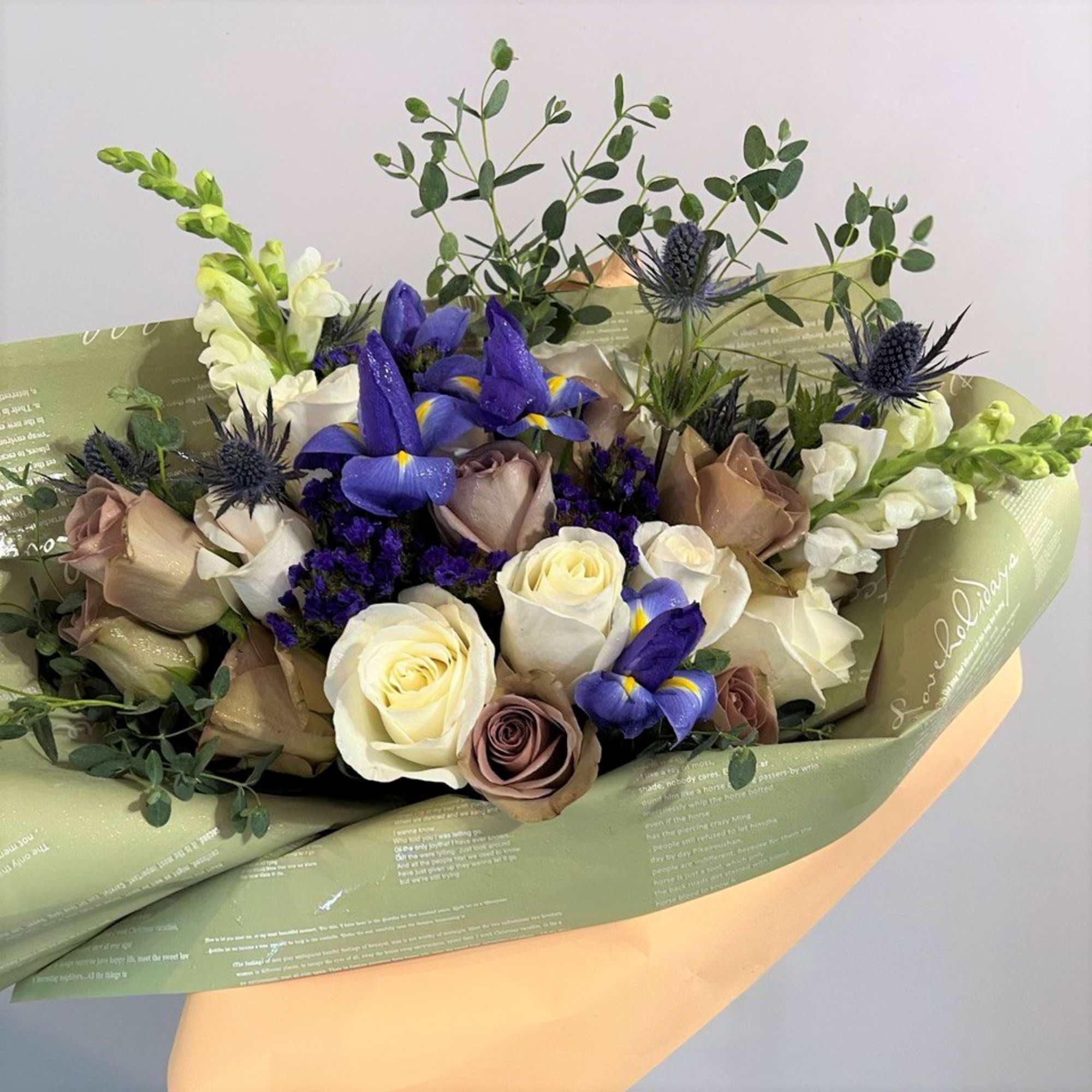 Hand-tied bouquet of white and taupe roses with purple irises and snapdragons in green printed wrap