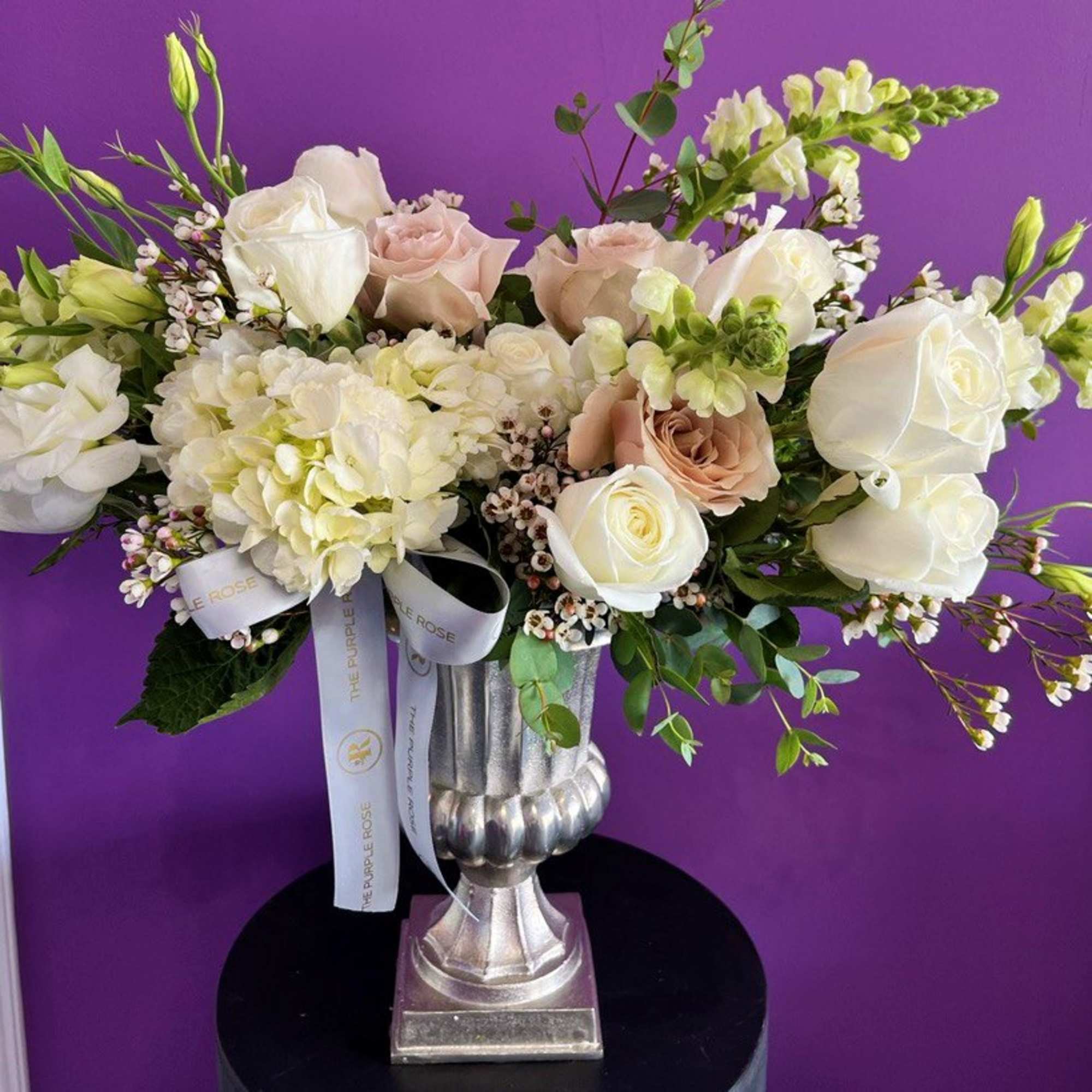Ivory and blush rose arrangement with hydrangea in a silver urn vase against a purple background