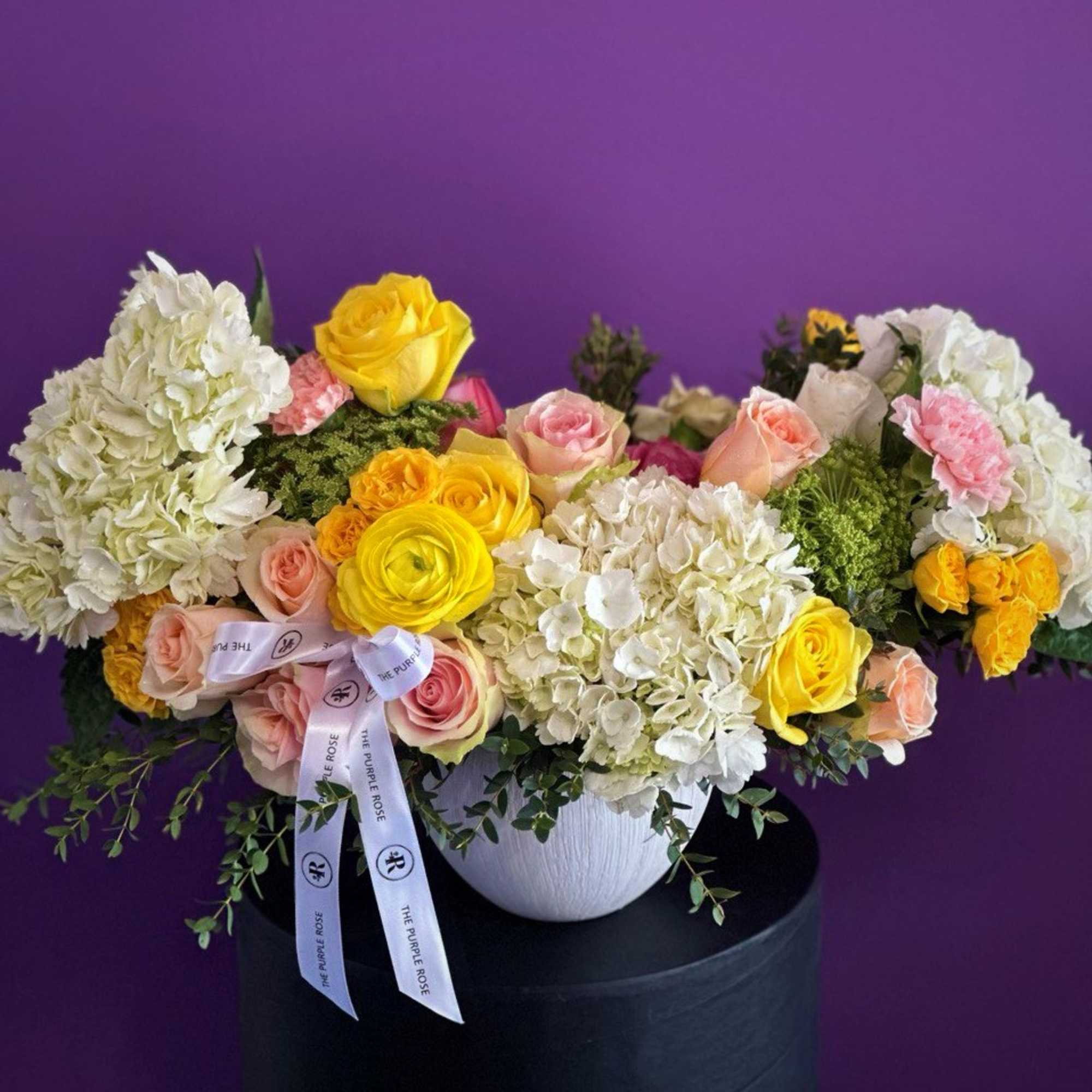 Low white bowl of white hydrangeas with yellow and peach roses against a purple background
