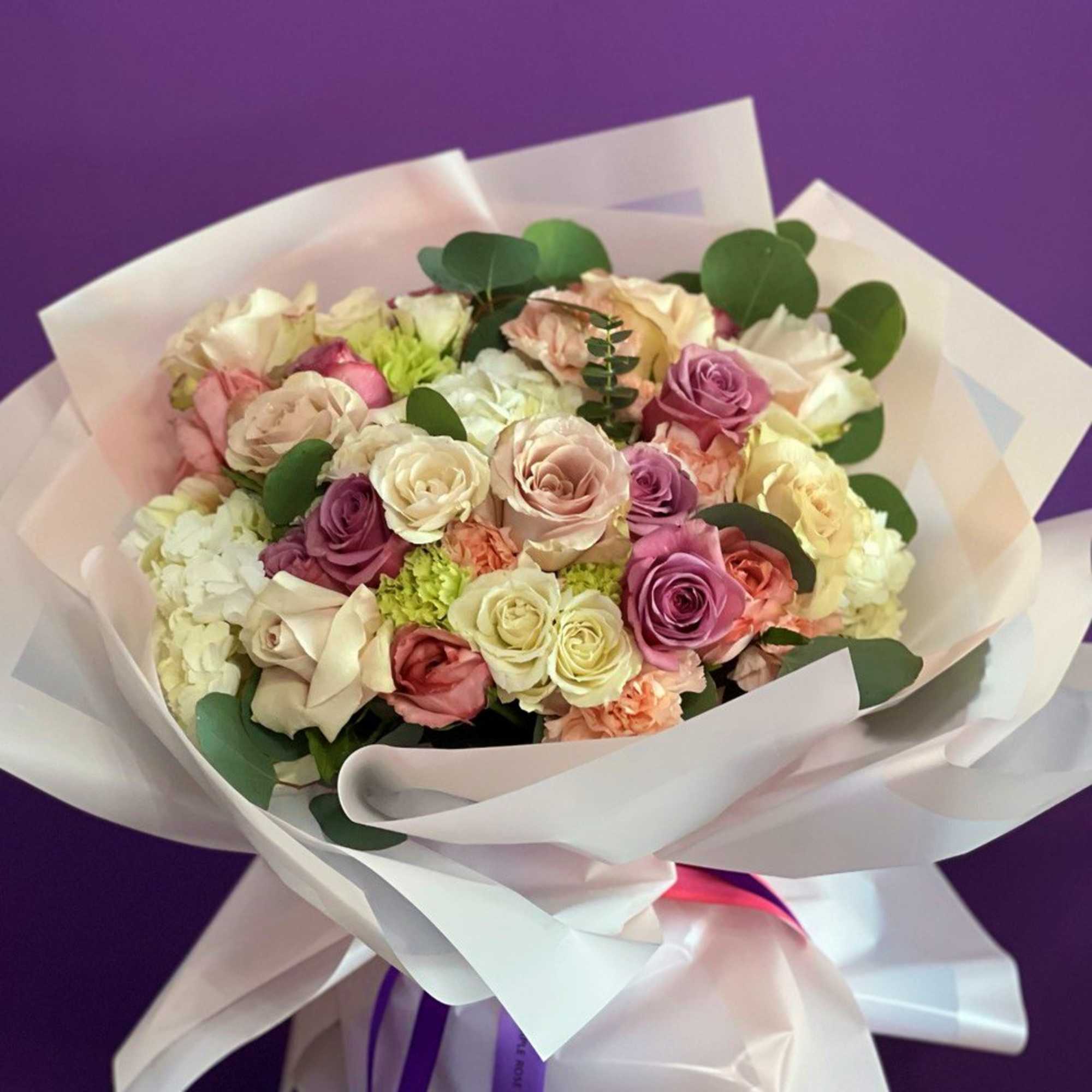 Hand-tied bouquet of pastel roses and white blooms wrapped in white paper.