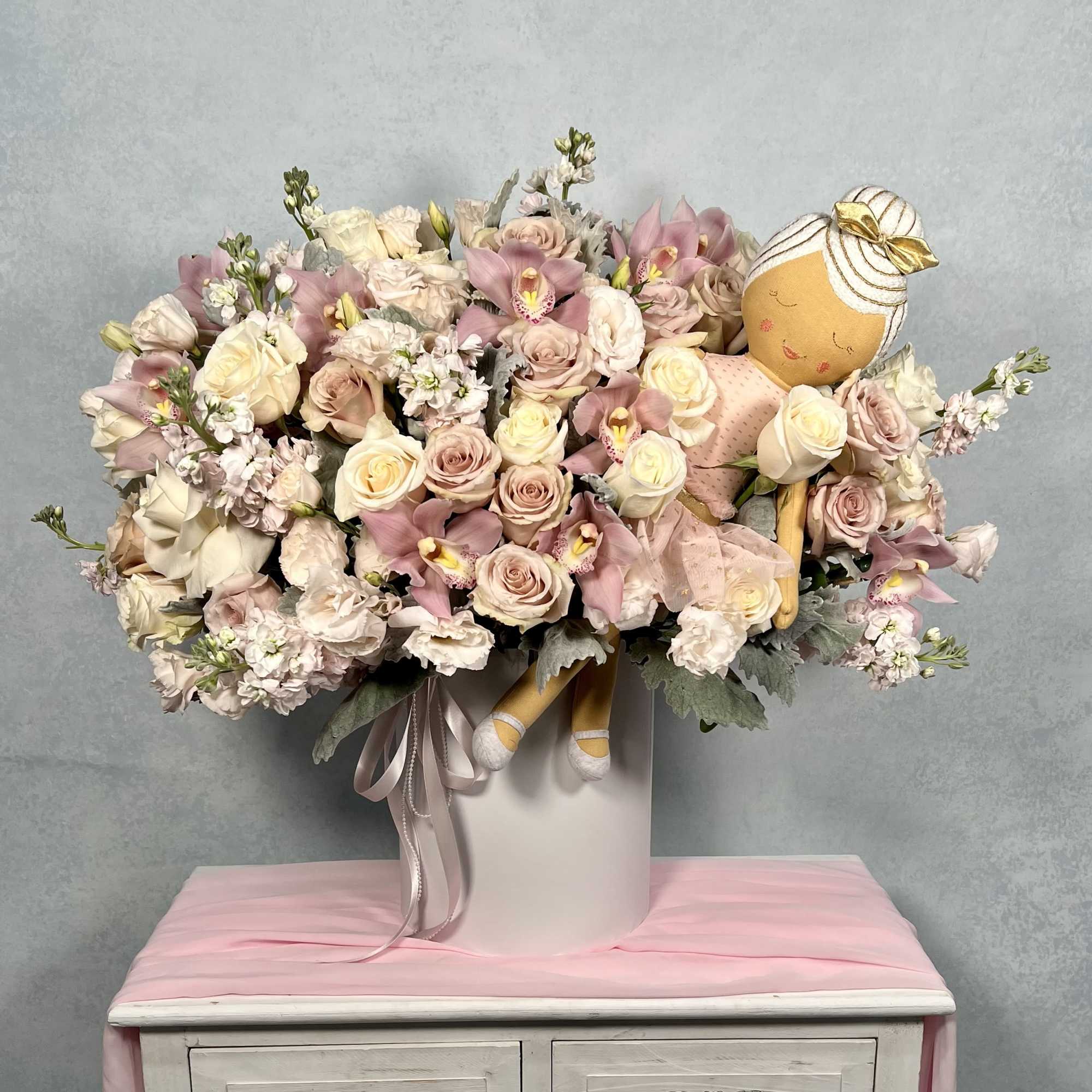 Large pastel pink and cream flower arrangement in a white hatbox with a plush ballerina doll nestled among the blooms