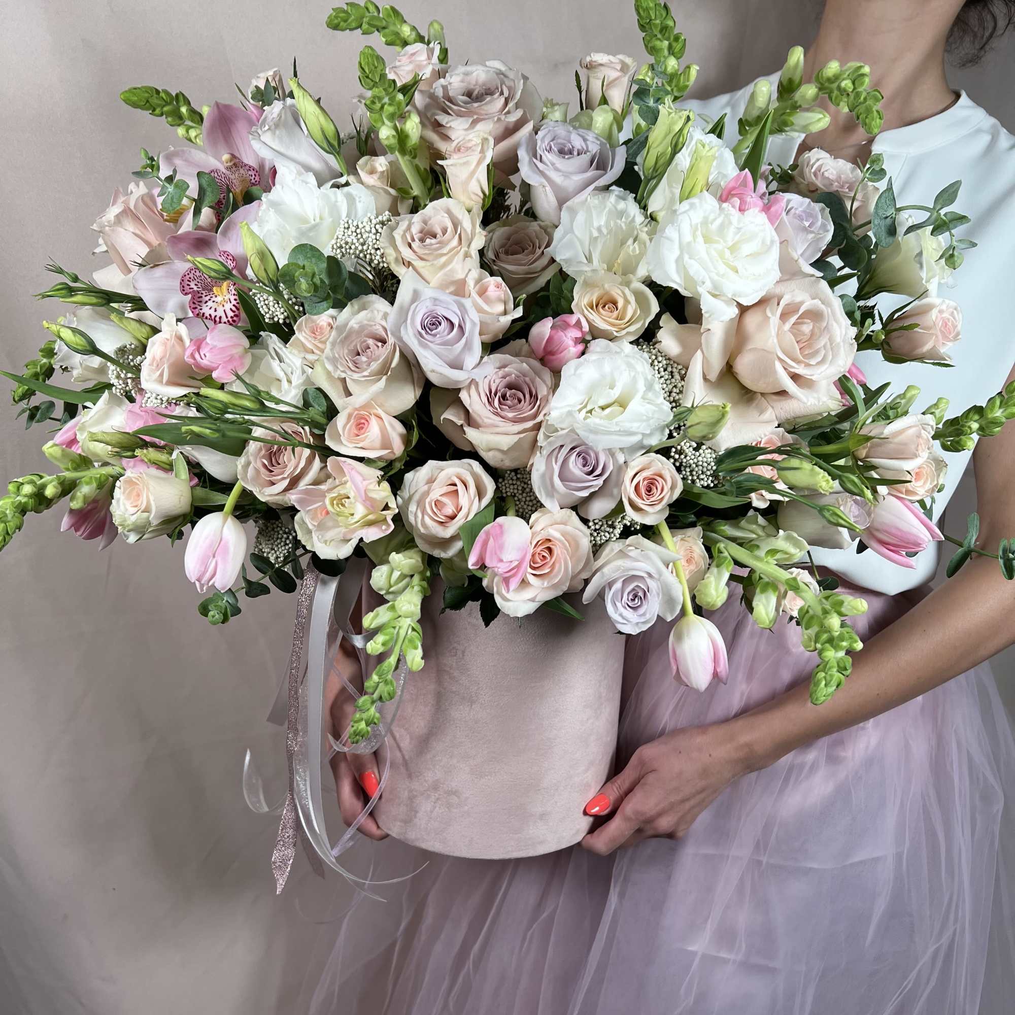 Large pastel arrangement of roses, tulips, and orchids in a blush hatbox held by a person