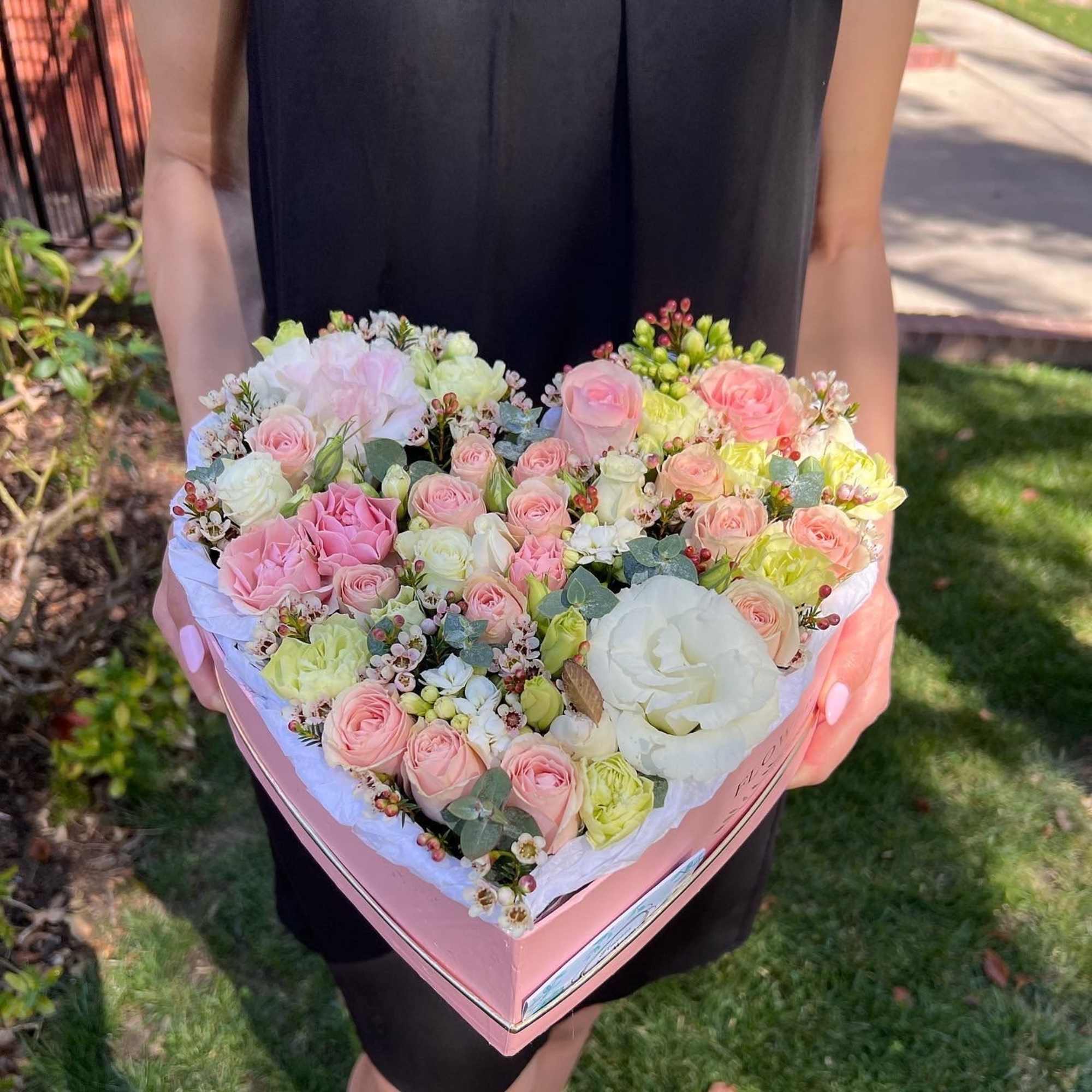 Heart-shaped pink box filled with pastel roses and carnations held by a person in a black dress.