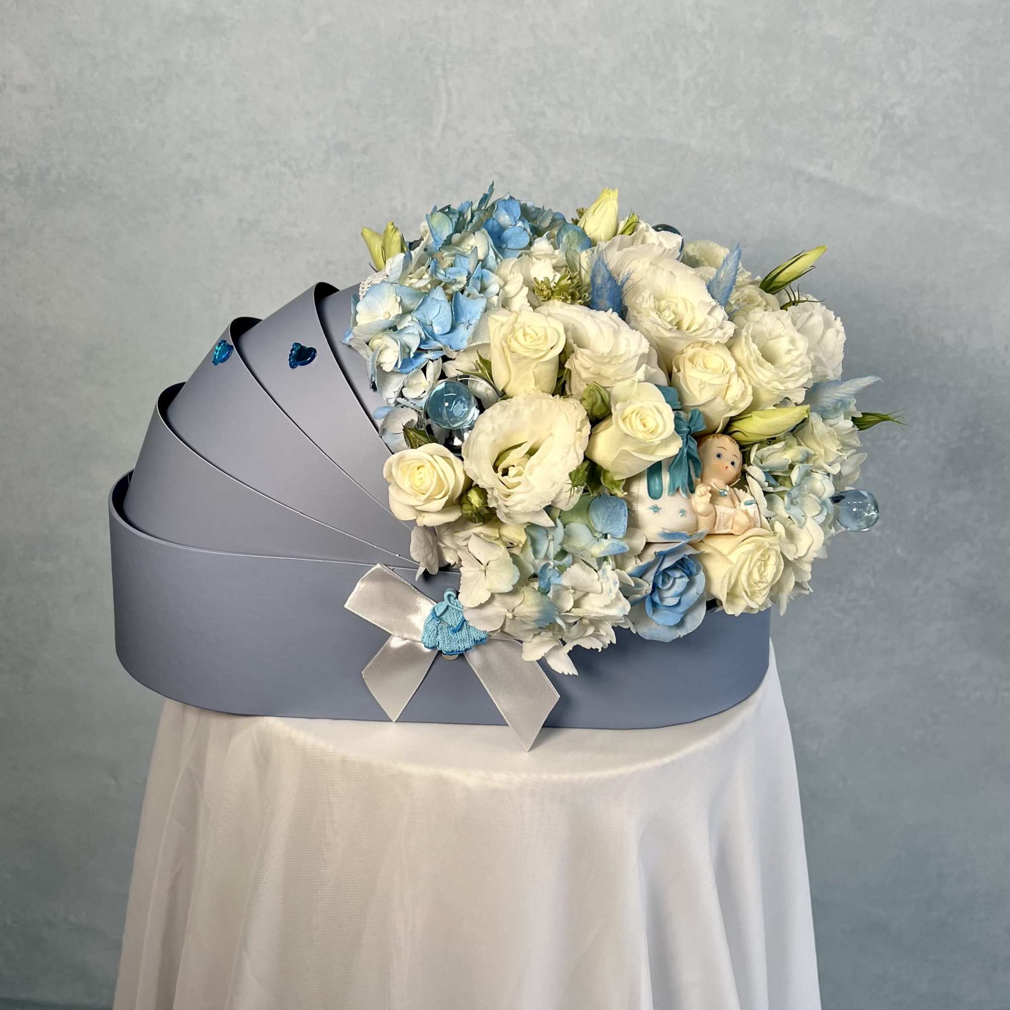 Light blue cradle-shaped box filled with white and blue flowers and a small baby figurine.