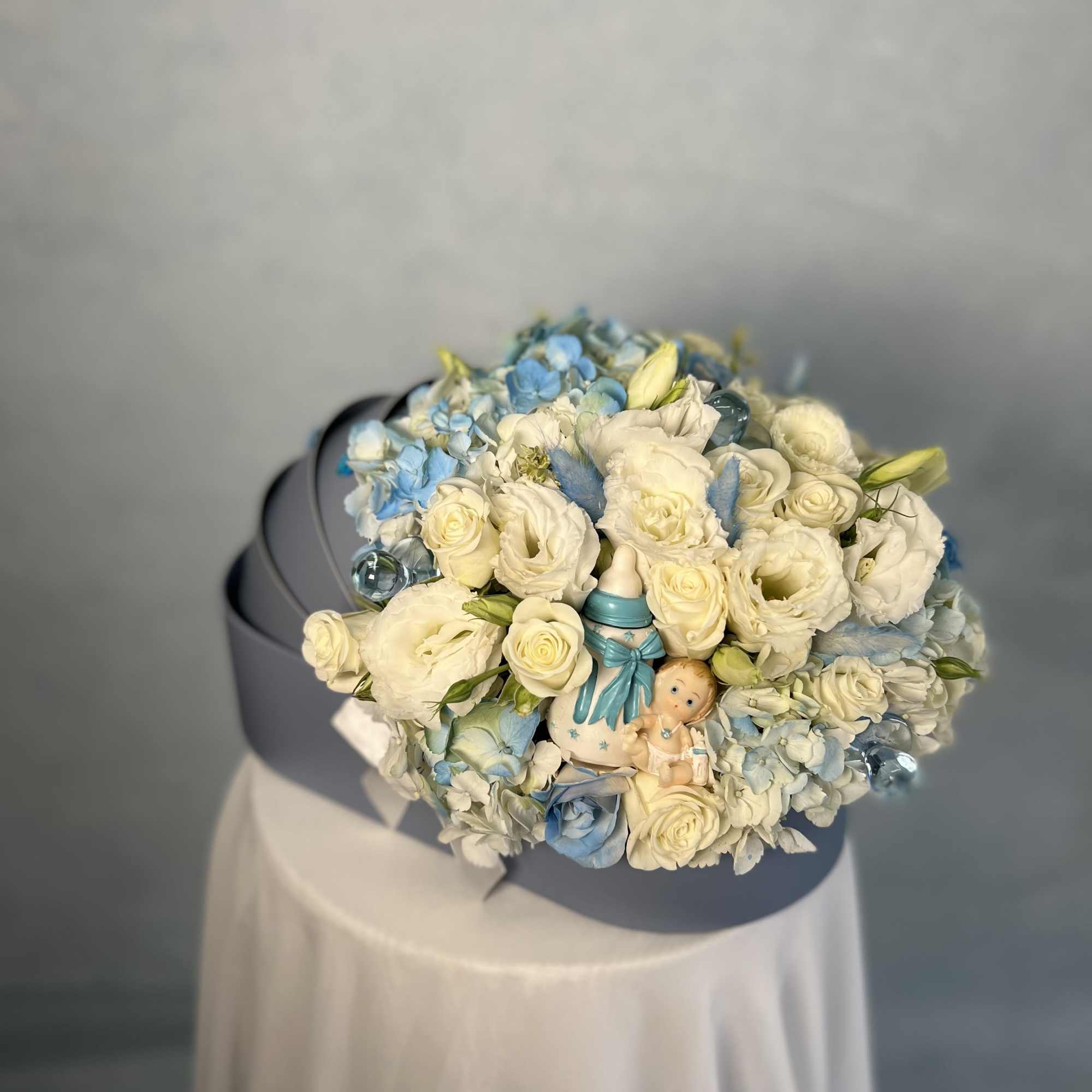 Round hat box arrangement of white and blue flowers with baby figurines and bottle decor.