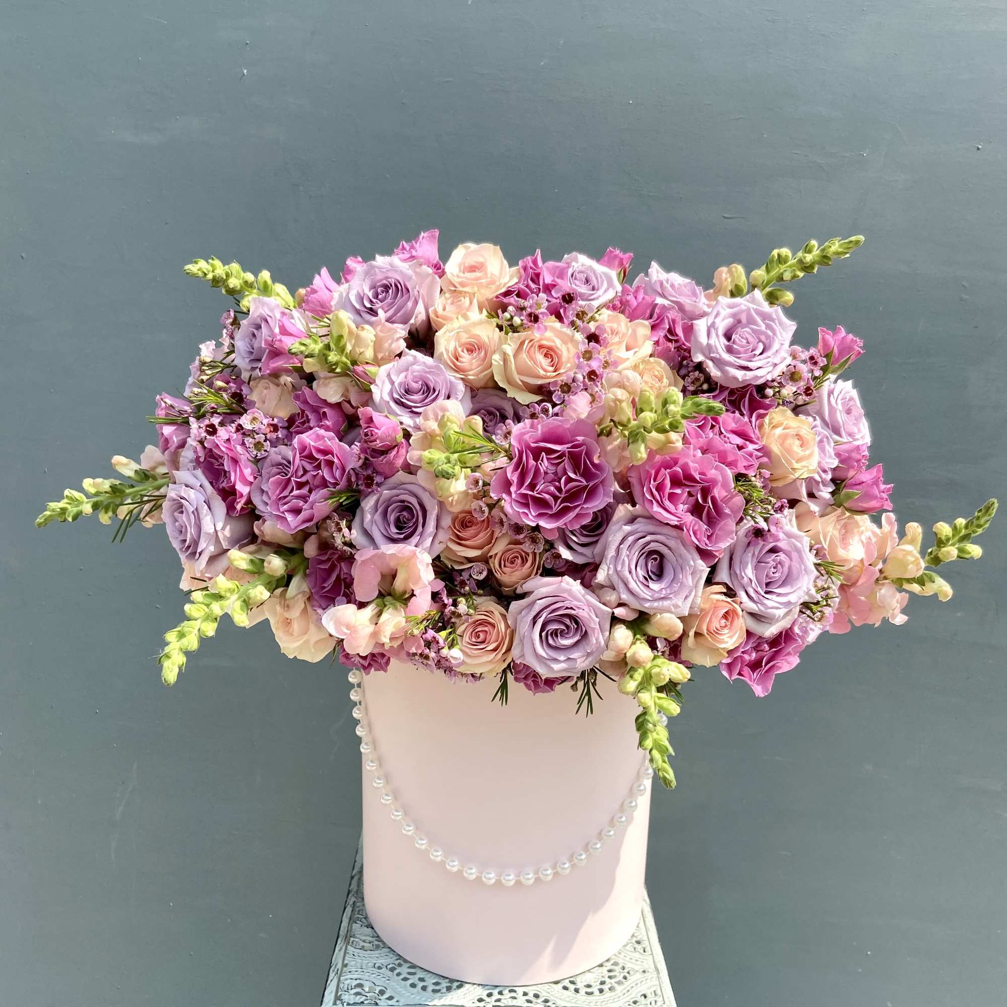 Lavender and pink rose arrangement in a pink hat box with pearls on a decorative metal stand.