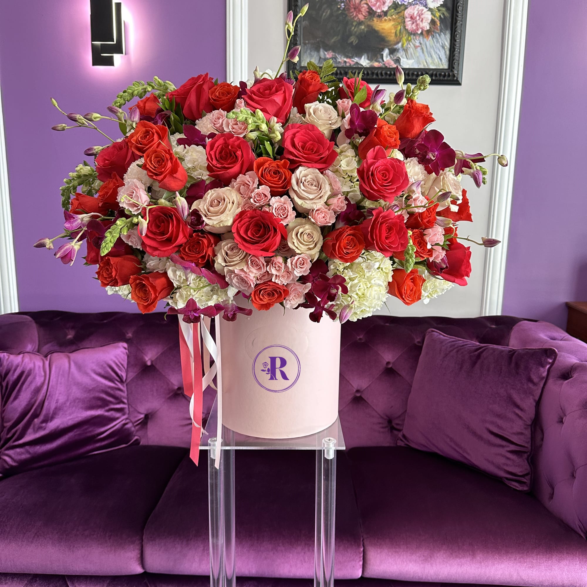 Large arrangement of red and pink roses with white blooms in a pink hatbox on a clear stand