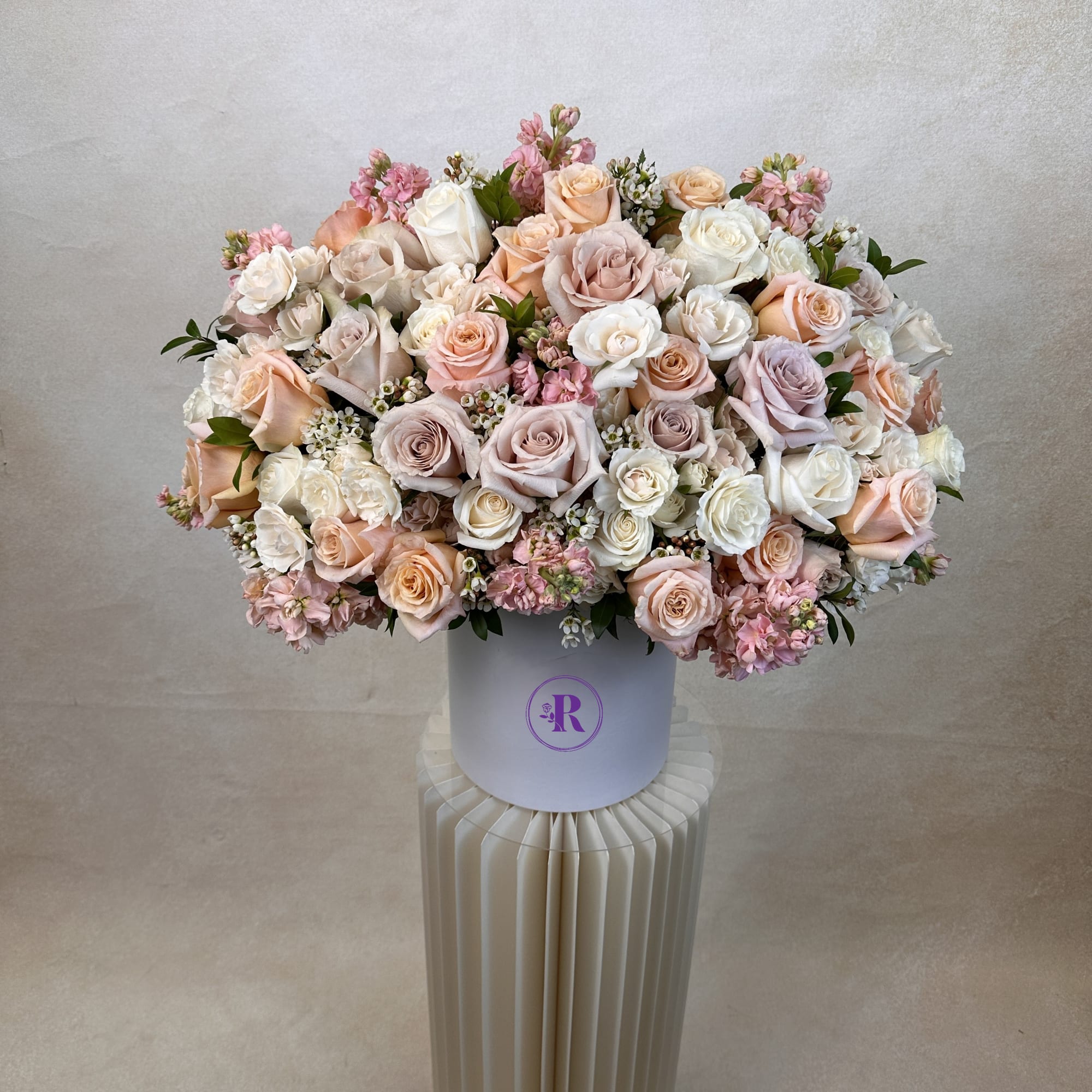 Round hatbox arrangement of pastel roses and small pink blooms on a pleated cream pedestal