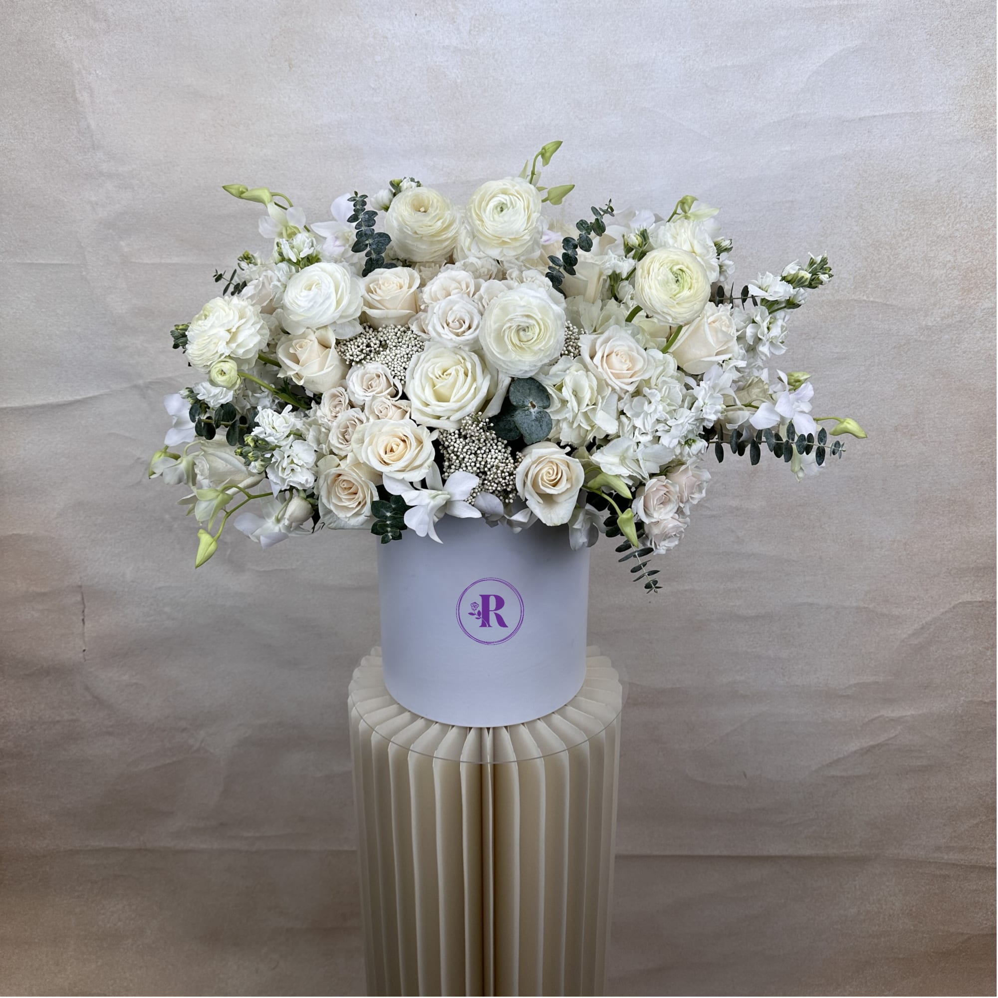 Large white and cream rose and ranunculus arrangement in a round white hatbox-style vase.