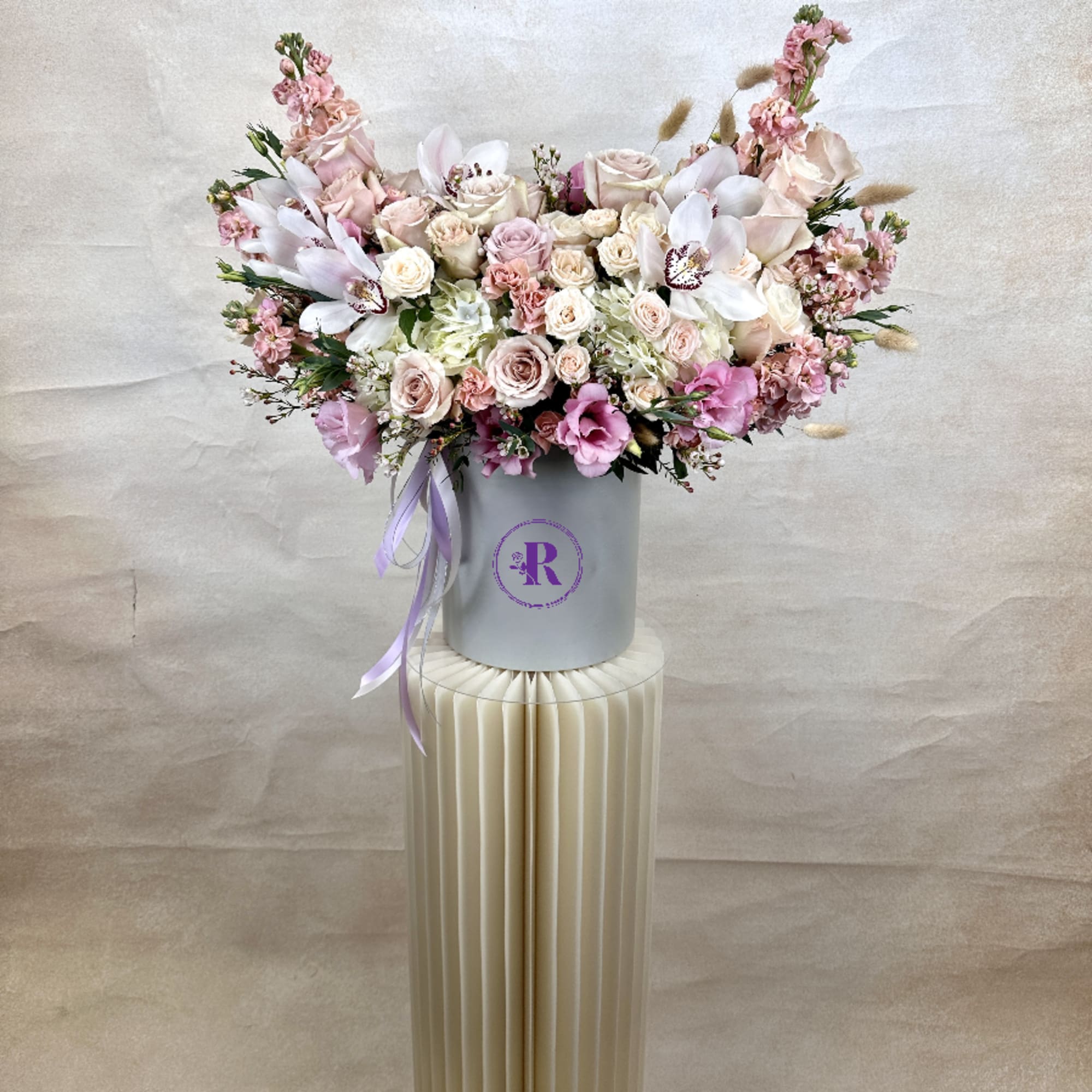 Wide pastel arrangement of roses, orchids, and mixed blooms in a white hatbox on a tall cream pedestal.