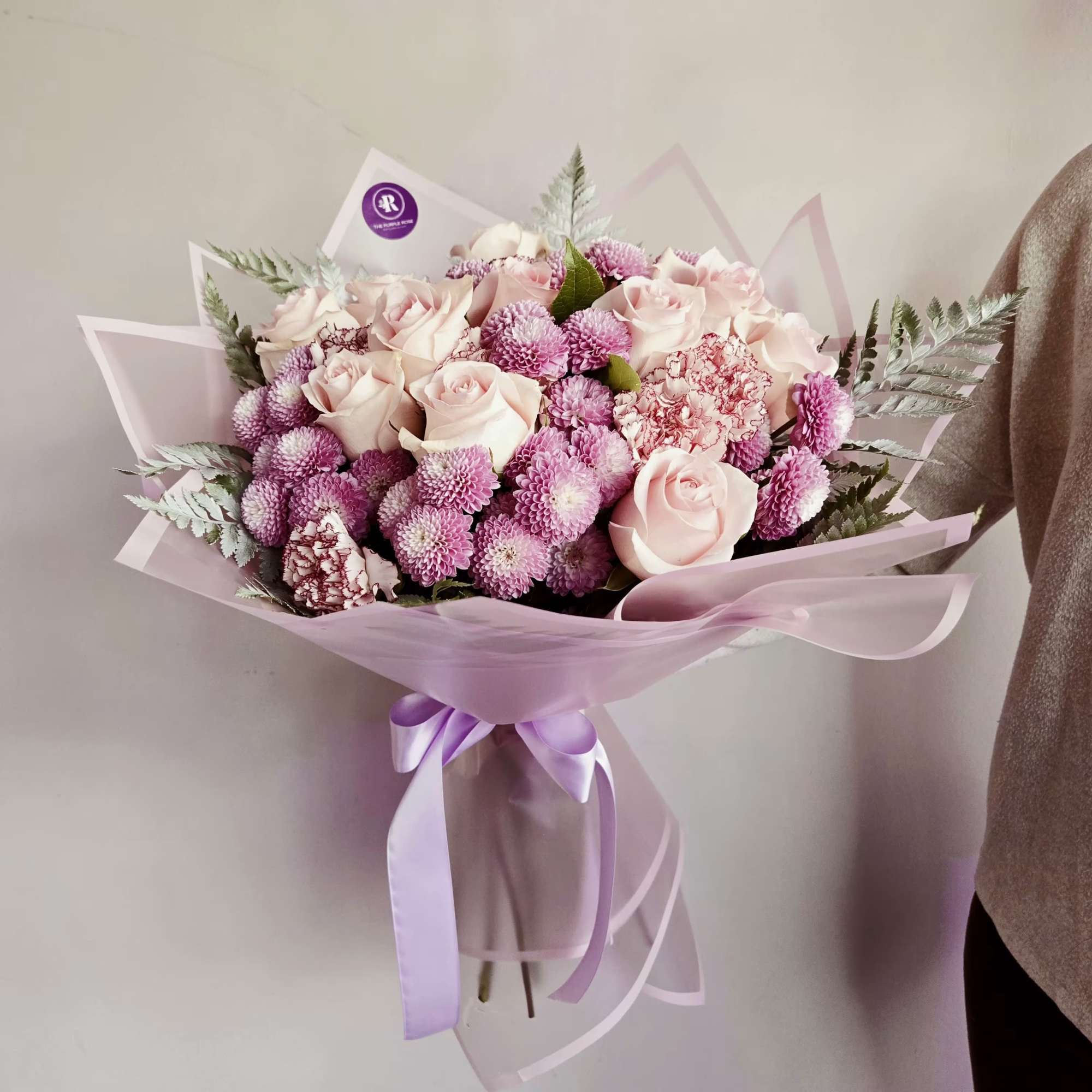 Handheld bouquet of pale pink roses and mauve pompom blooms wrapped in pink paper with a lavender ribbon.