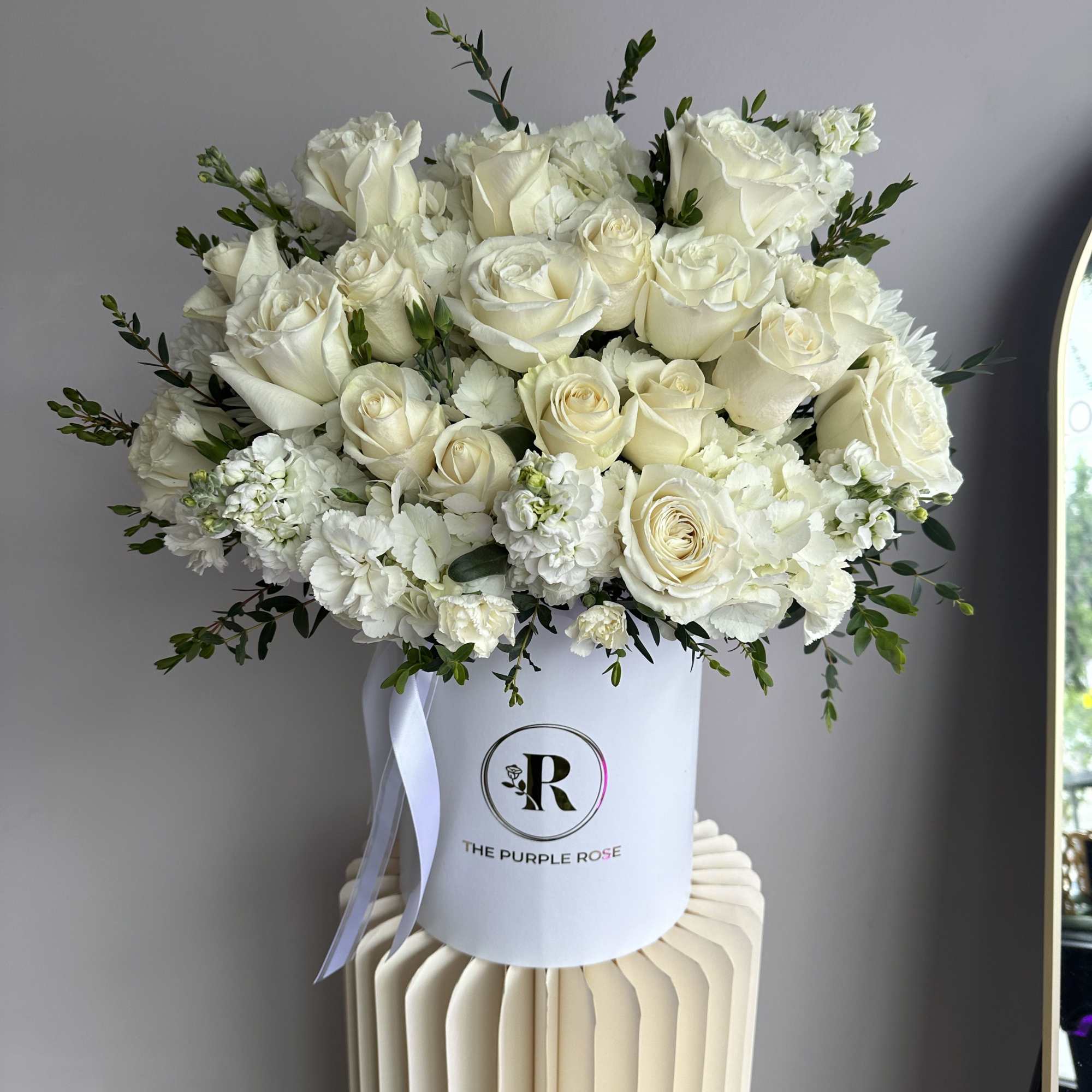 Round hatbox of white roses and mixed white flowers with greenery on a cream pedestal
