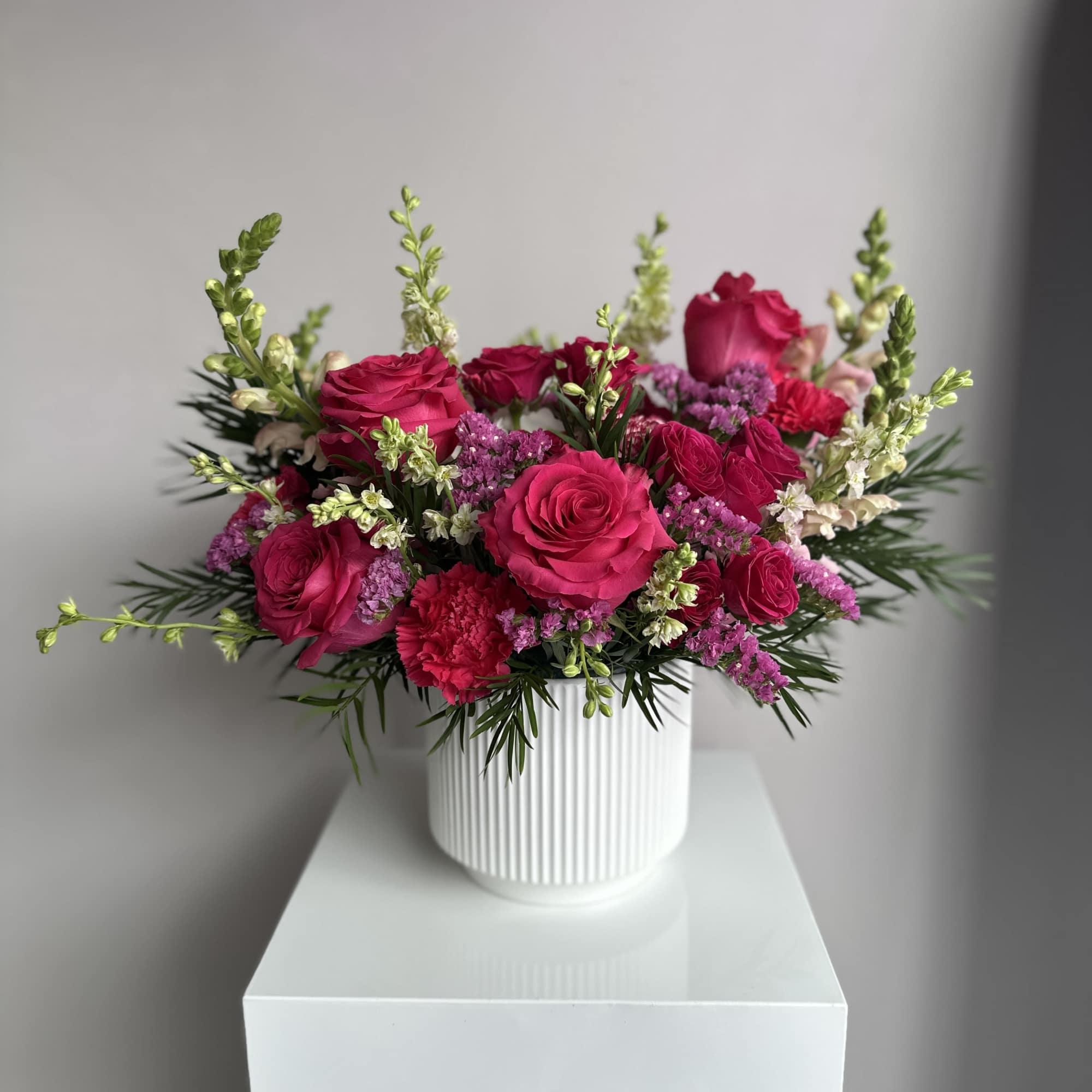 Bright pink roses and carnations arranged with cream blooms in a white ribbed ceramic vase.