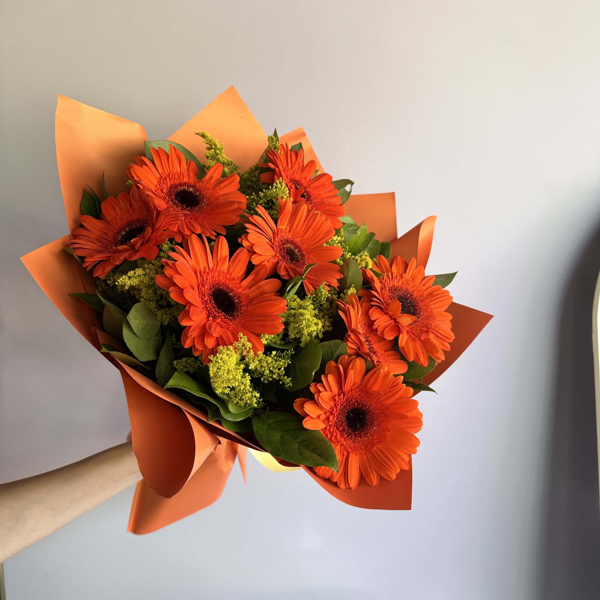 Handheld bouquet of bright orange gerbera daisies with yellow filler flowers in orange wrap.