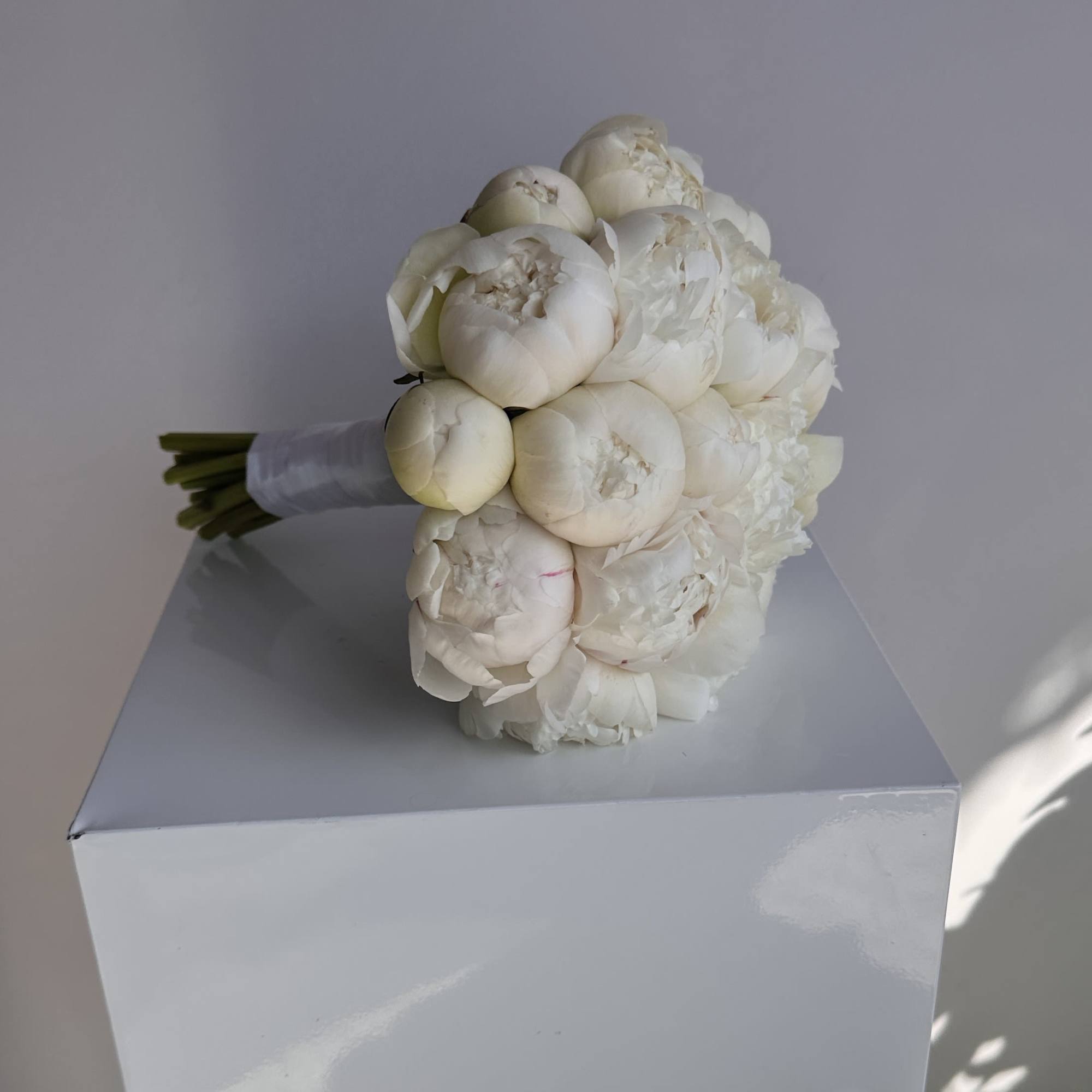 Compact white peony hand-tied bouquet with wrapped stems on a white pedestal.