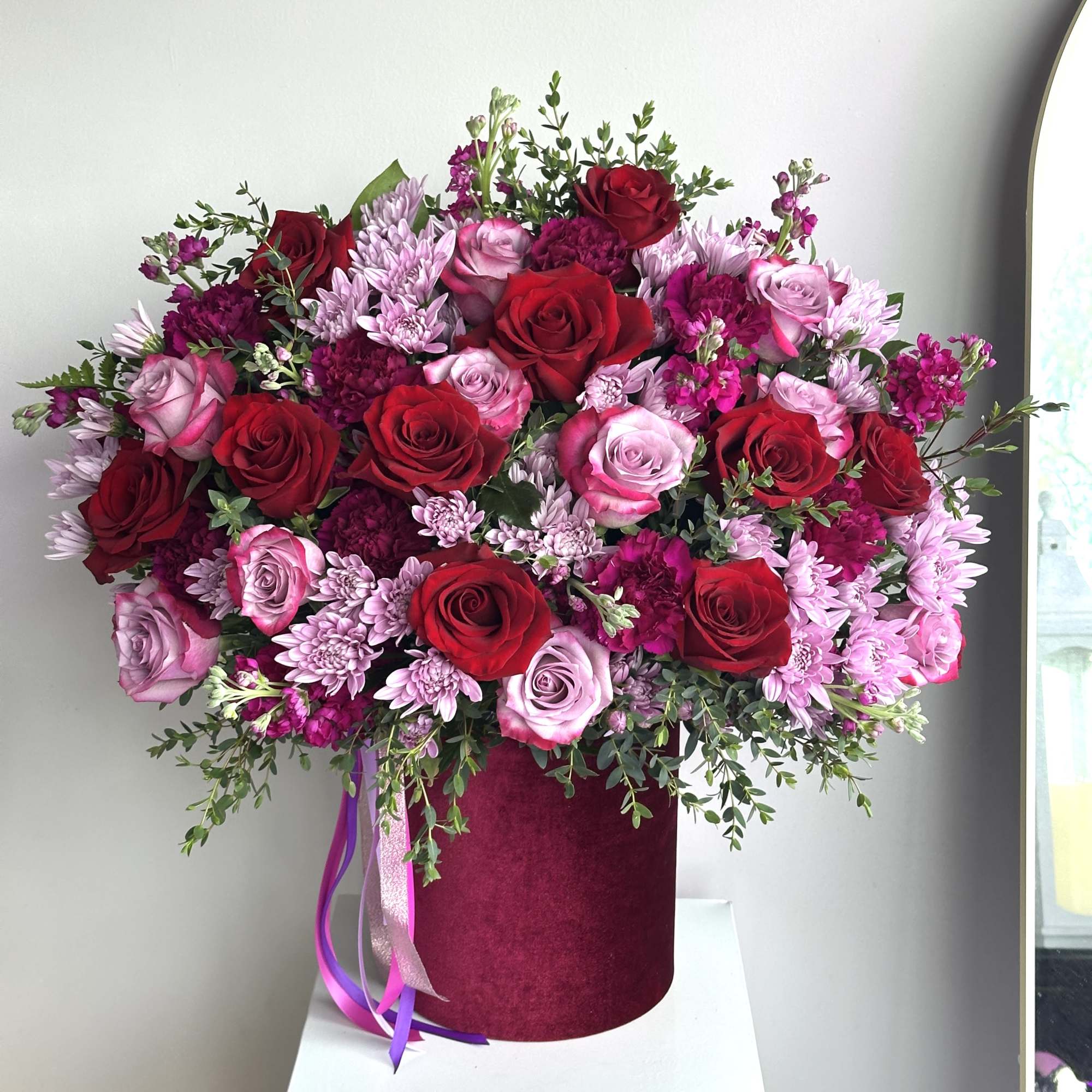 Large arrangement of red and pink roses with pink mums in a dark pink hatbox with ribbon