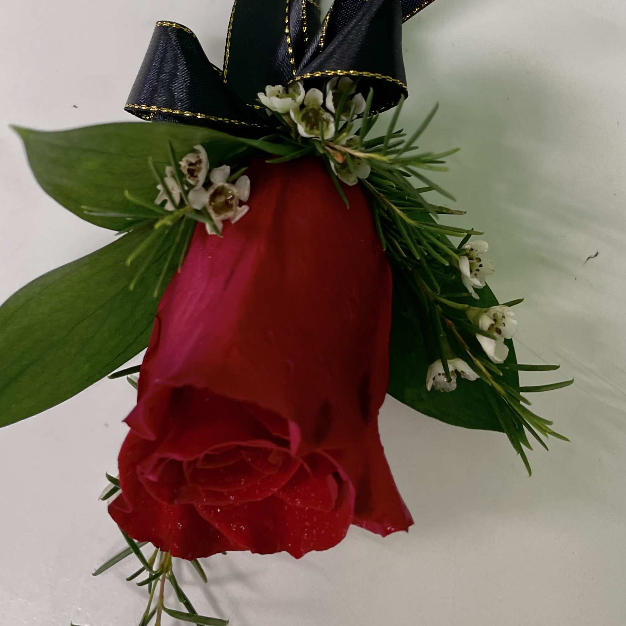 Single red rose with greenery, small white blossoms, and a black ribbon bow