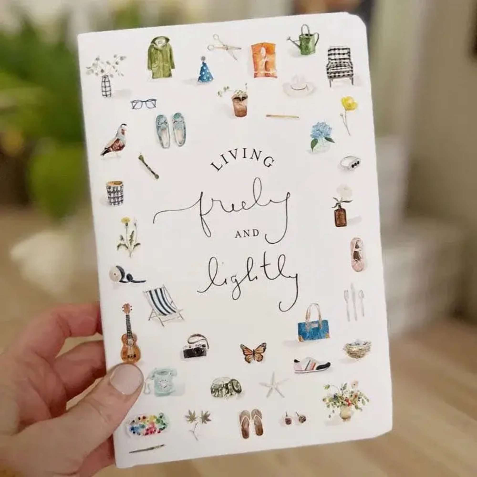 Hand holding a white notebook with small illustrated objects and the words "living freely and lightly"