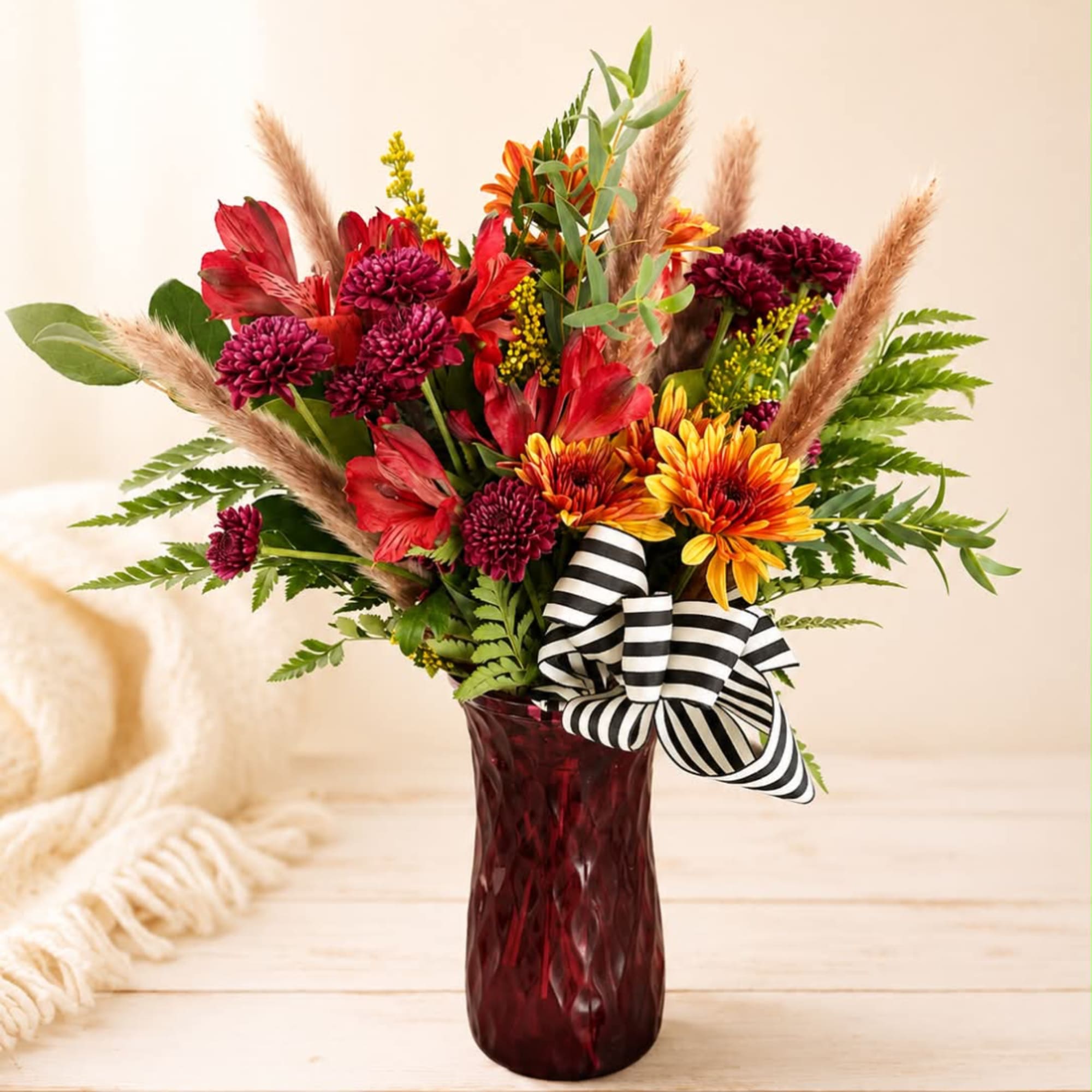 Mixed bouquet of red and orange flowers in a burgundy vase with a striped ribbon