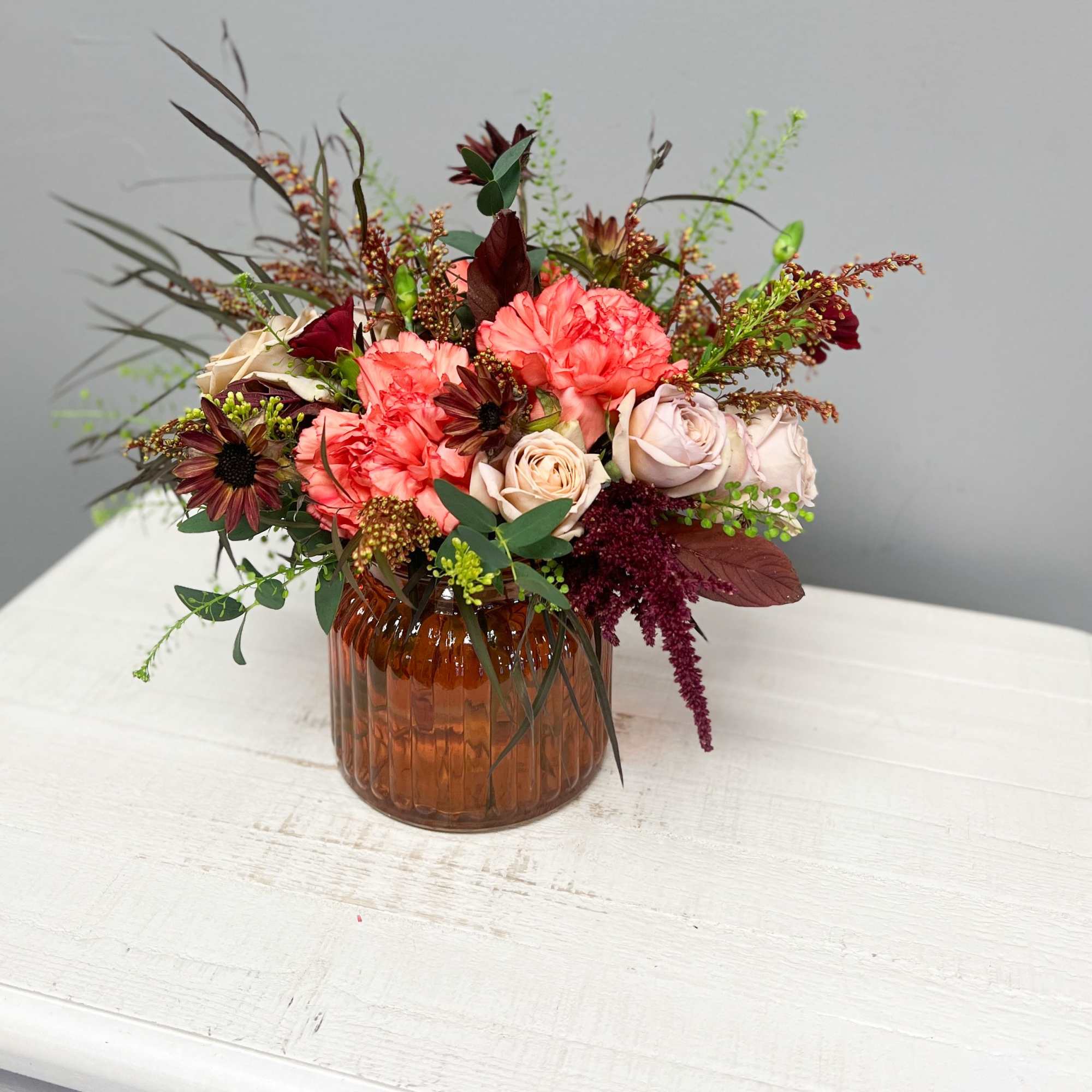 Mixed bouquet of coral, blush, and burgundy flowers in an amber glass vase