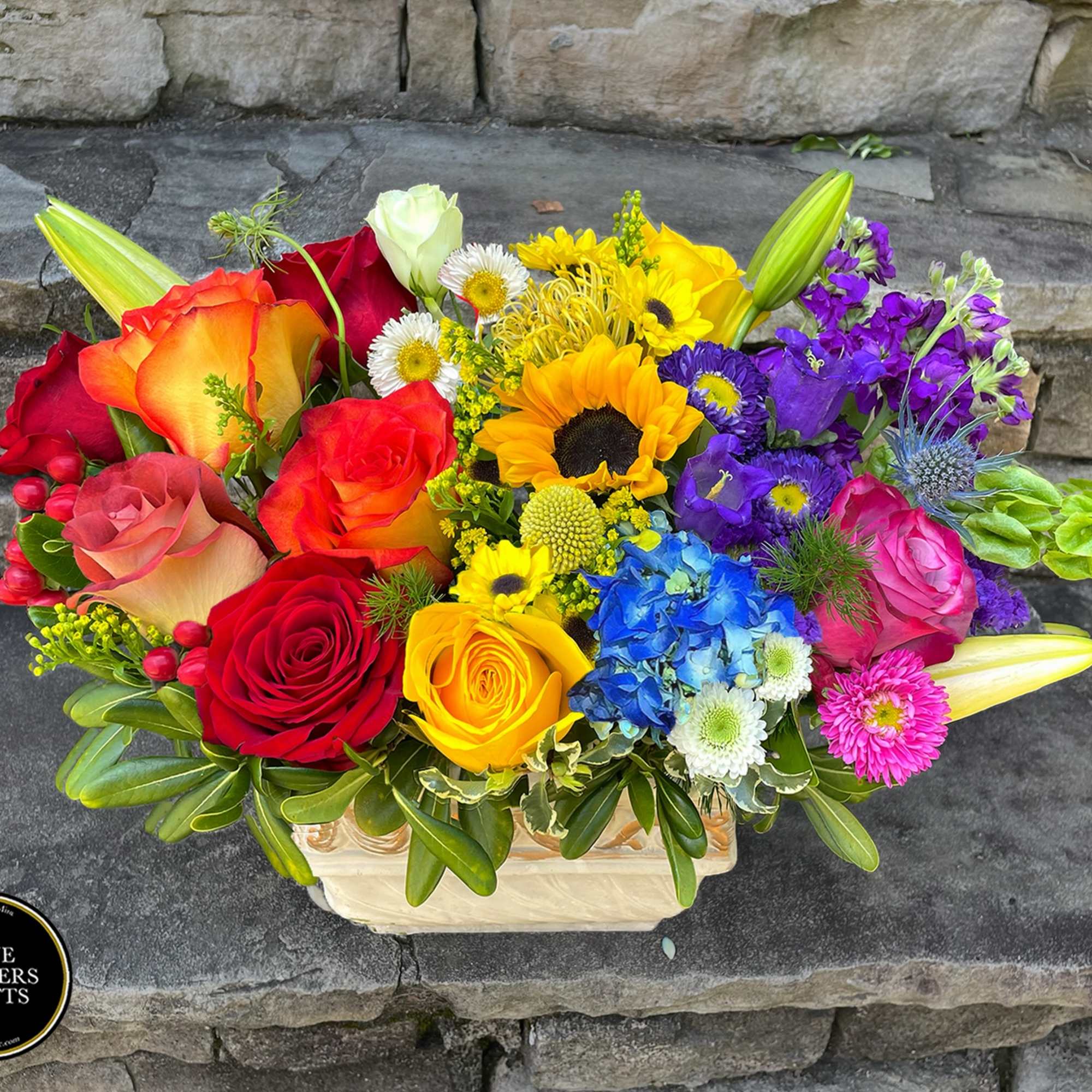 Low wooden box arrangement of multicolored roses, sunflower, hydrangea, lilies, and other bright flowers.