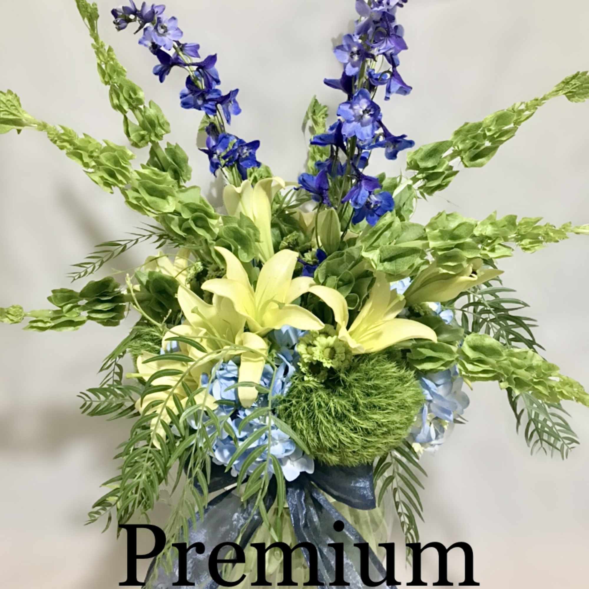 Tall arrangement of blue delphinium, green bells of Ireland, yellow lilies, and blue hydrangeas in a glass vase