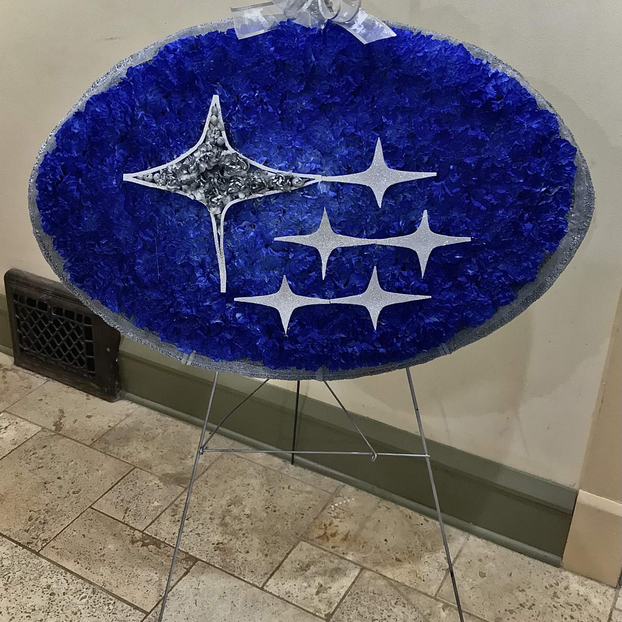 Oval blue floral display with silver star shapes on an easel, topped with a sheer silver bow.