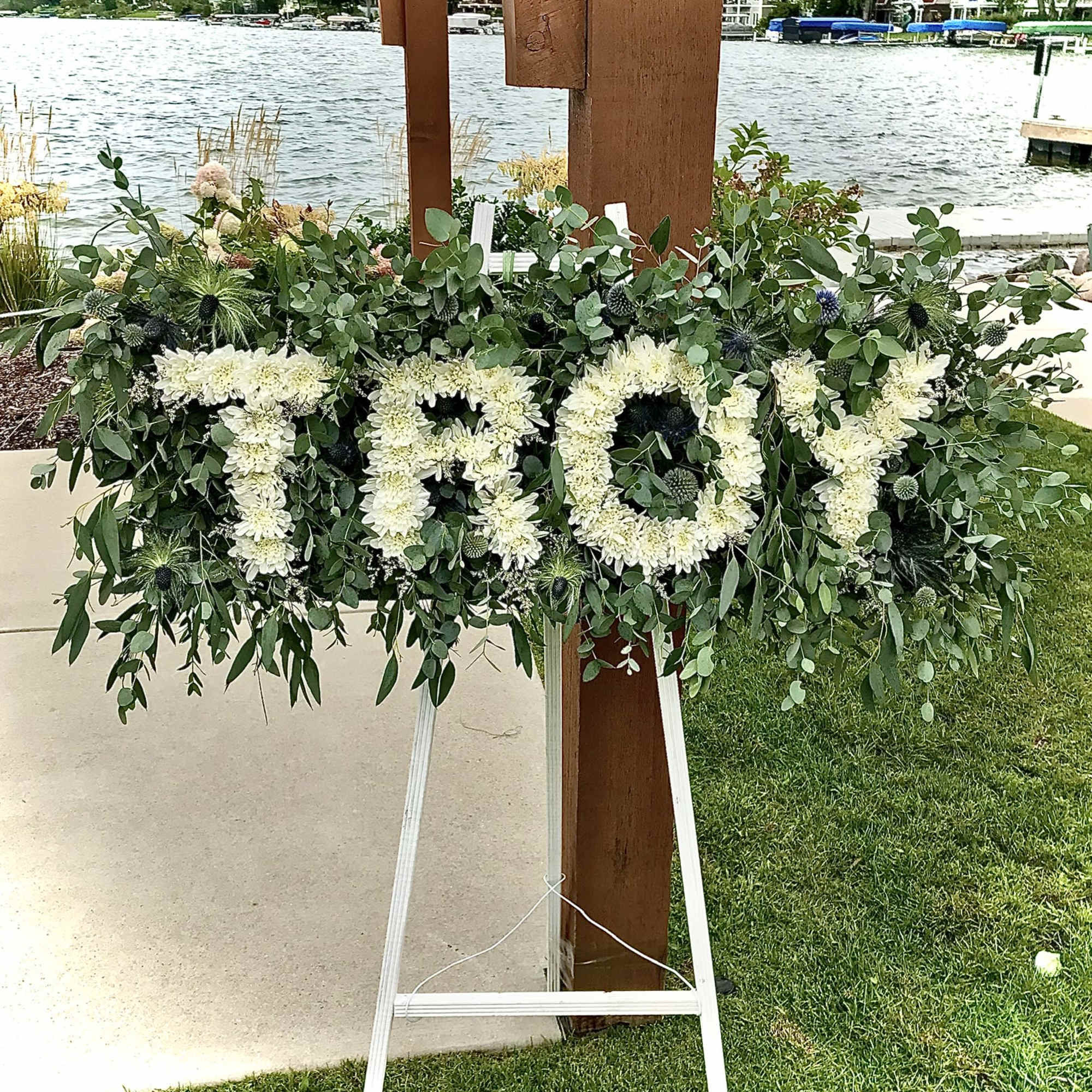 Standing floral easel spelling TROY in white flowers against dense greenery by a lakeside