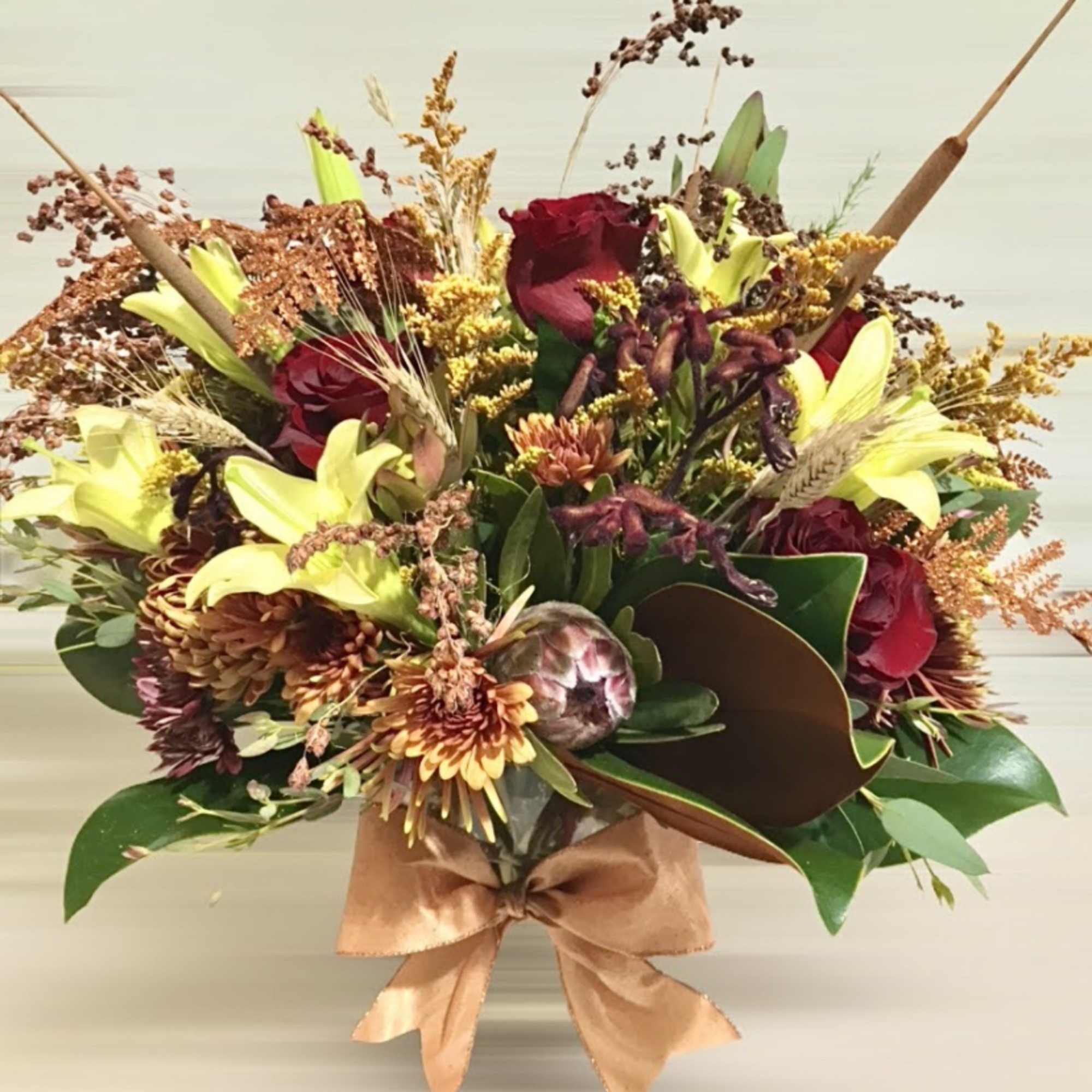 Autumn bouquet with red roses, yellow lilies, and bronze mums in a vase with a tan bow.