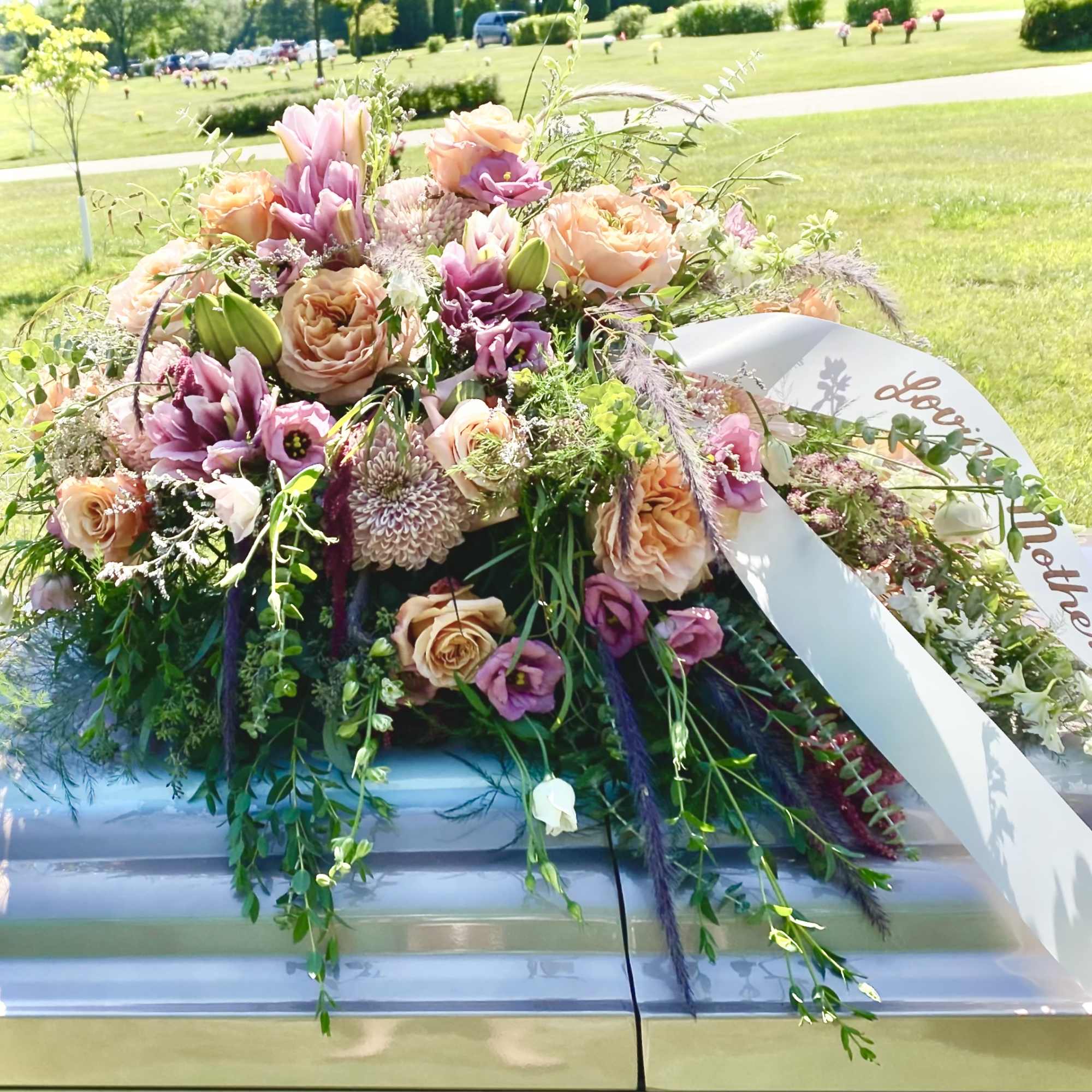 Peach and mauve casket spray with roses and lilies on a silver casket with memorial ribbon